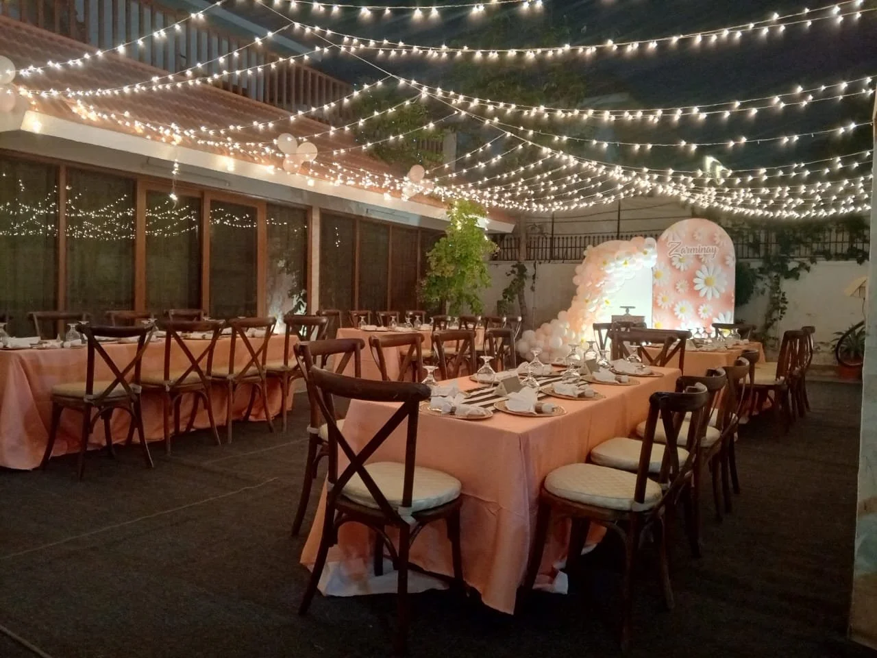 Decorated outdoor dining area for a celebration with string lights, balloons, and a floral arch backdrop.