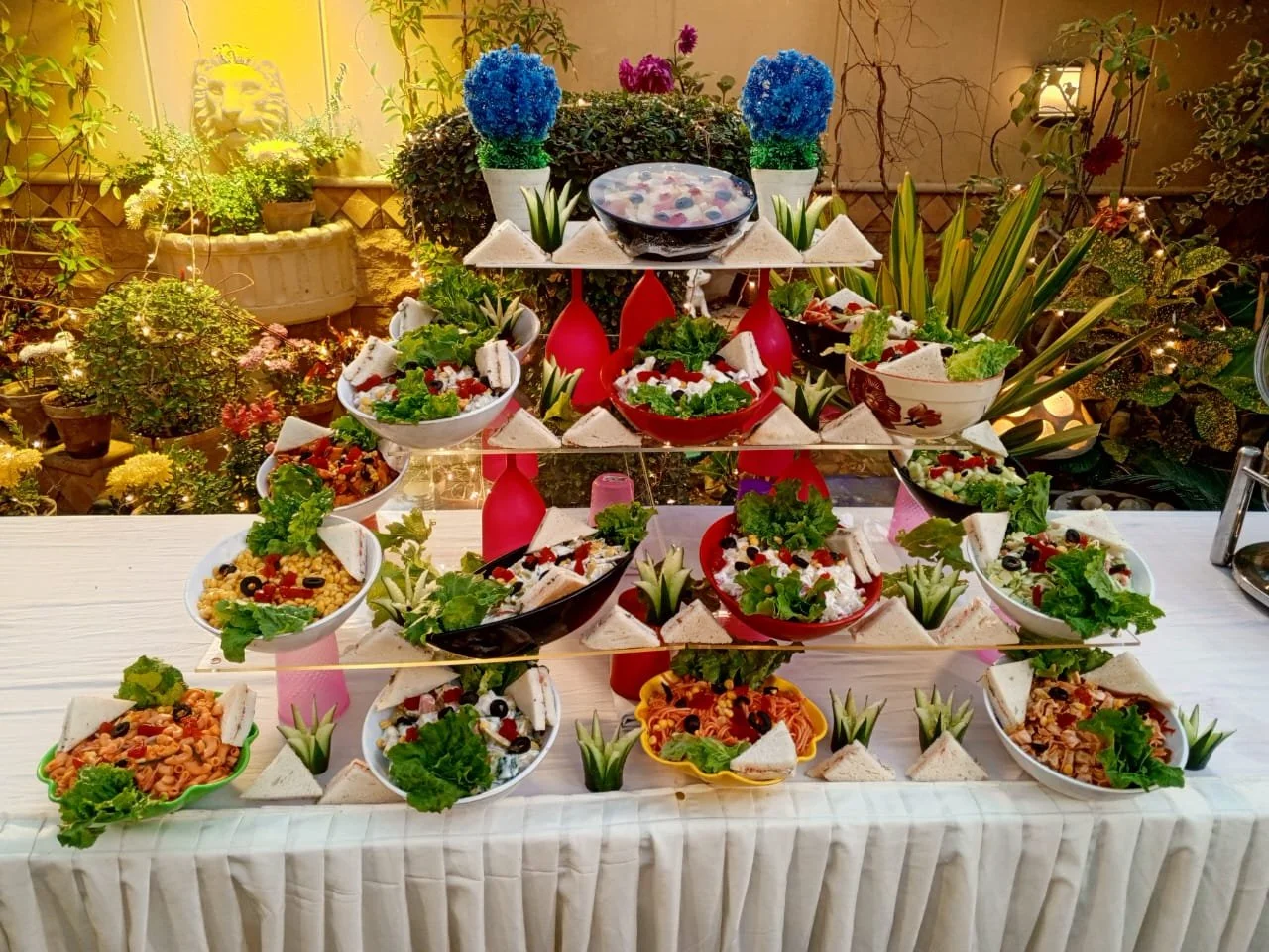 Tiered display of various colorful salads and appetizers on a table, decorated with small plant arrangements, with potted plants and greenery in the background.