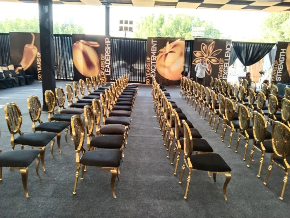 Rows of black and gold chairs arranged in an event space with large banners on the back wall displaying gold and peach-colored apples and motivational words, with people setting up or preparing for an event.