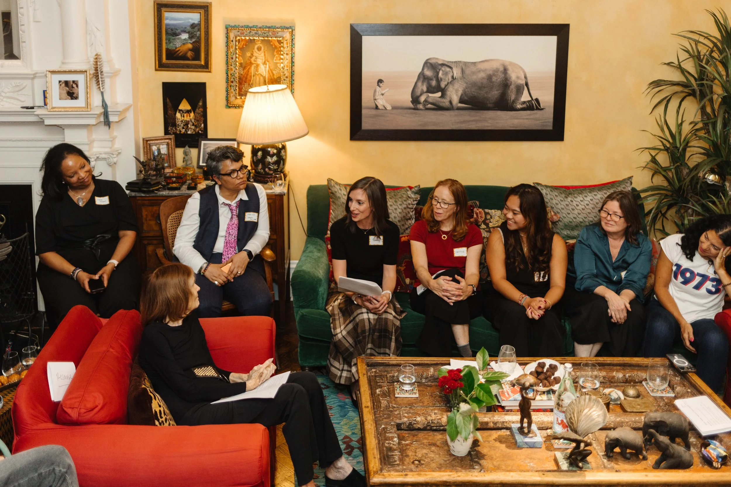 A diverse group of women are sitting and having a discussion in a cozy, well-decorated living room, with artwork on the walls and a wooden coffee table with decorations.