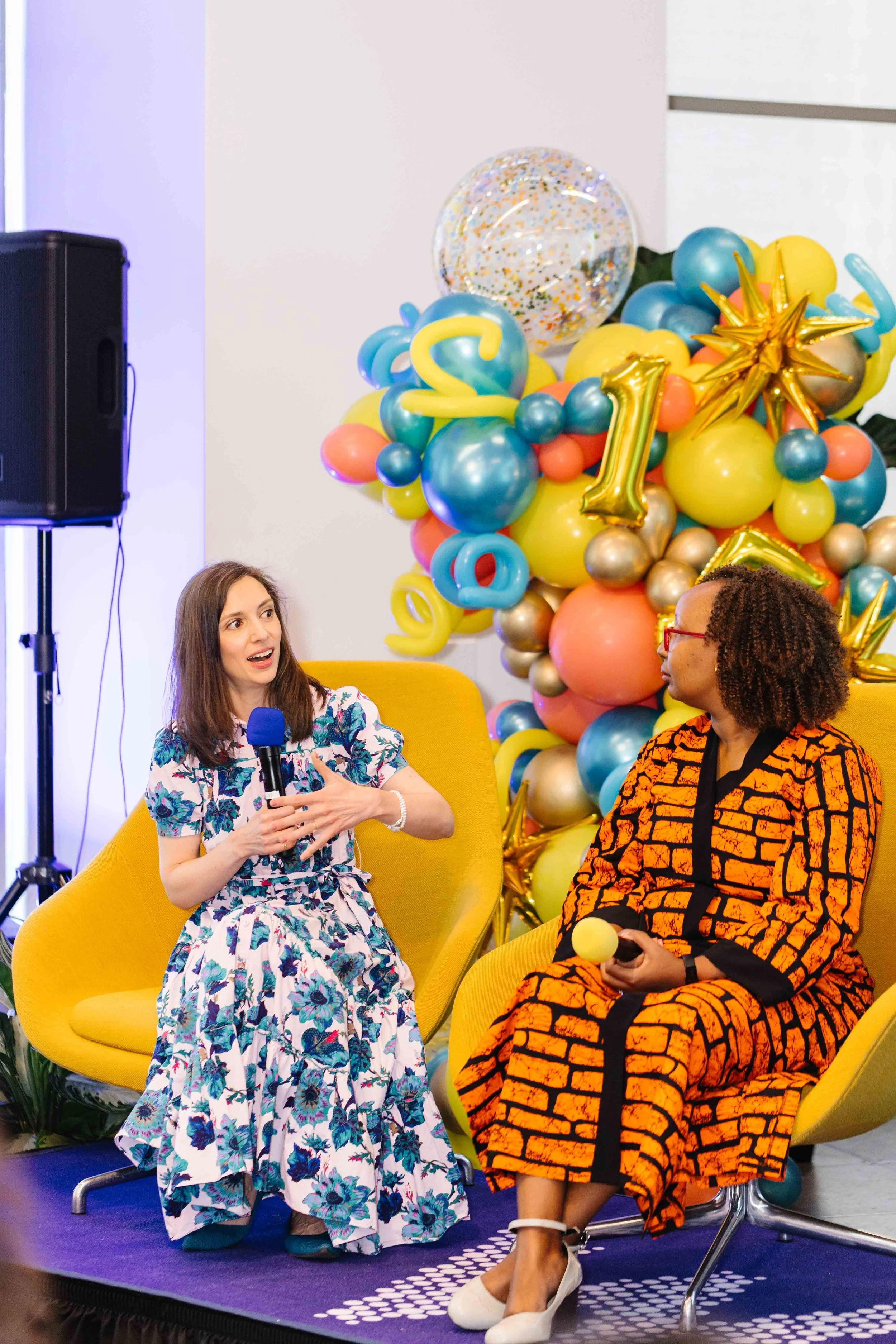 Two women seated on yellow chairs in front of colorful balloon decorations, one speaking holding a blue microphone, and the other holding a yellow microphone at an indoor event.