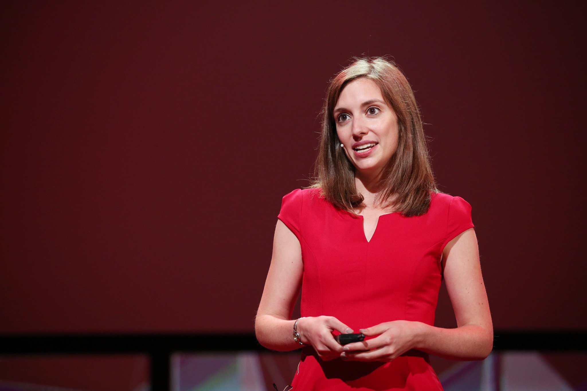 A woman with shoulder-length brown hair wearing a red dress is speaking on stage, holding a remote control or phone in her hands, against a dark red background.