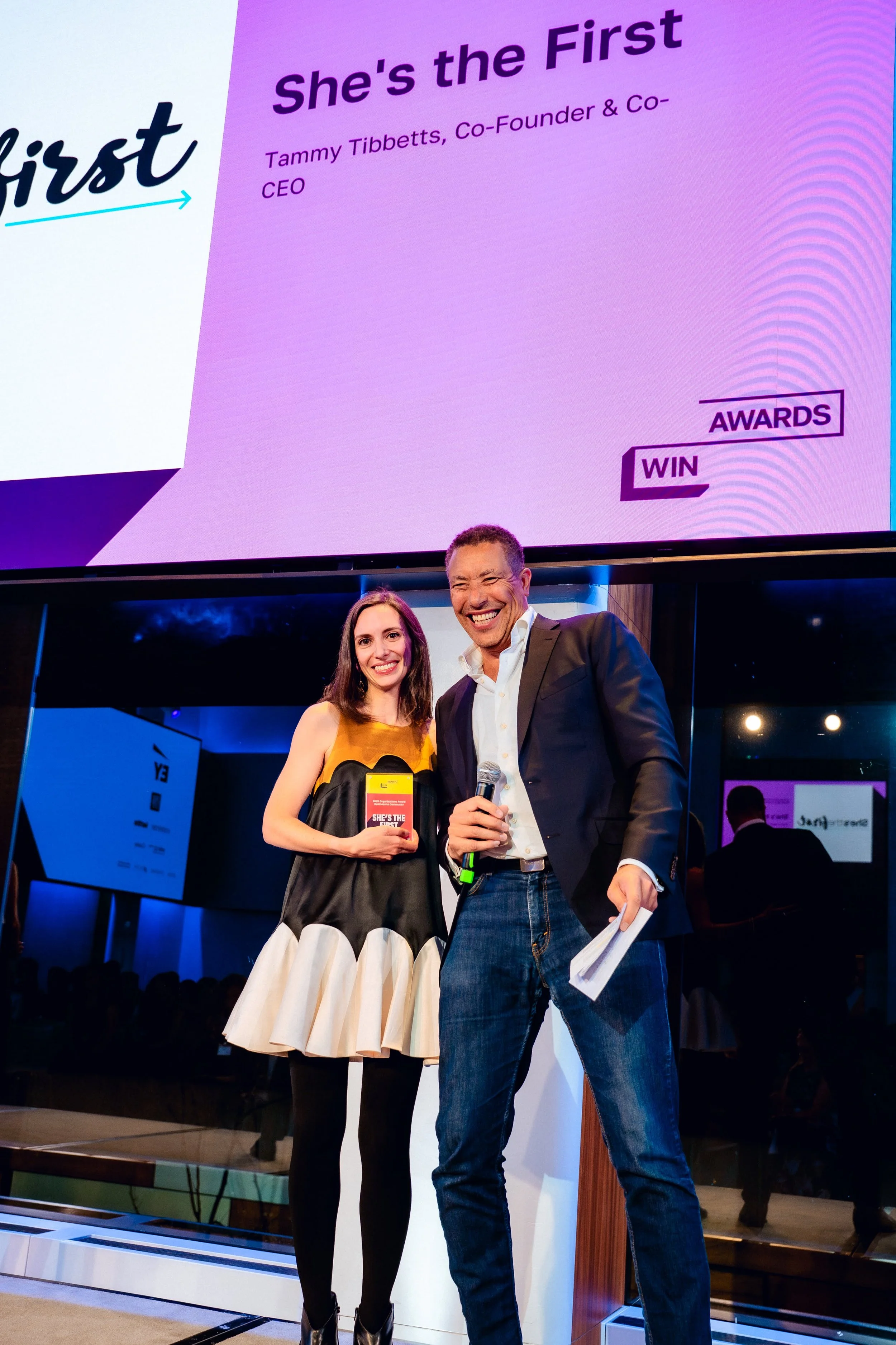 Woman in a sleeveless dress holding an award, standing next to a man in a suit with a microphone, smiling on stage at an awards ceremony.