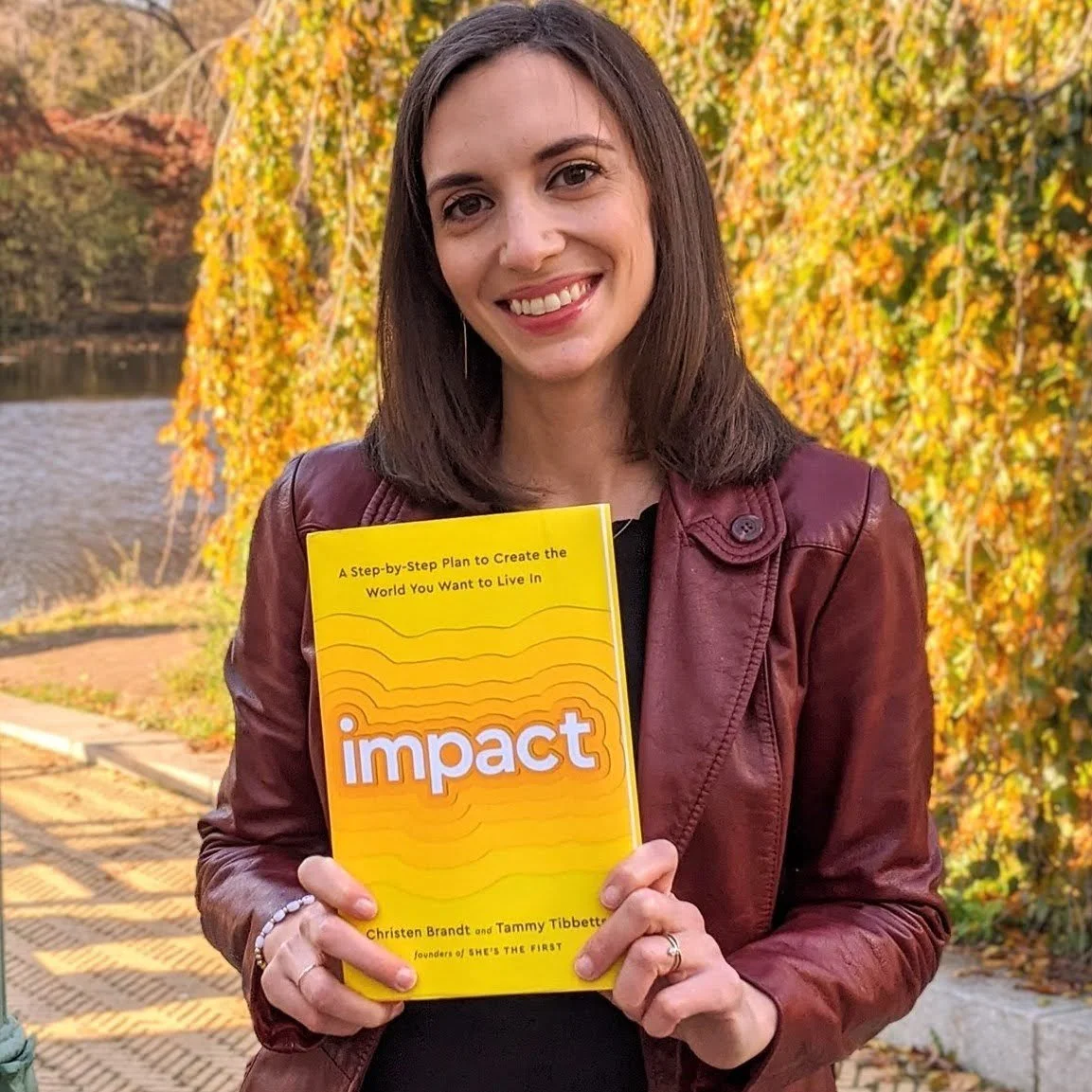 A woman with shoulder-length brown hair smiling outdoors, holding a yellow book titled 'Impact' in front of autumn foliage.