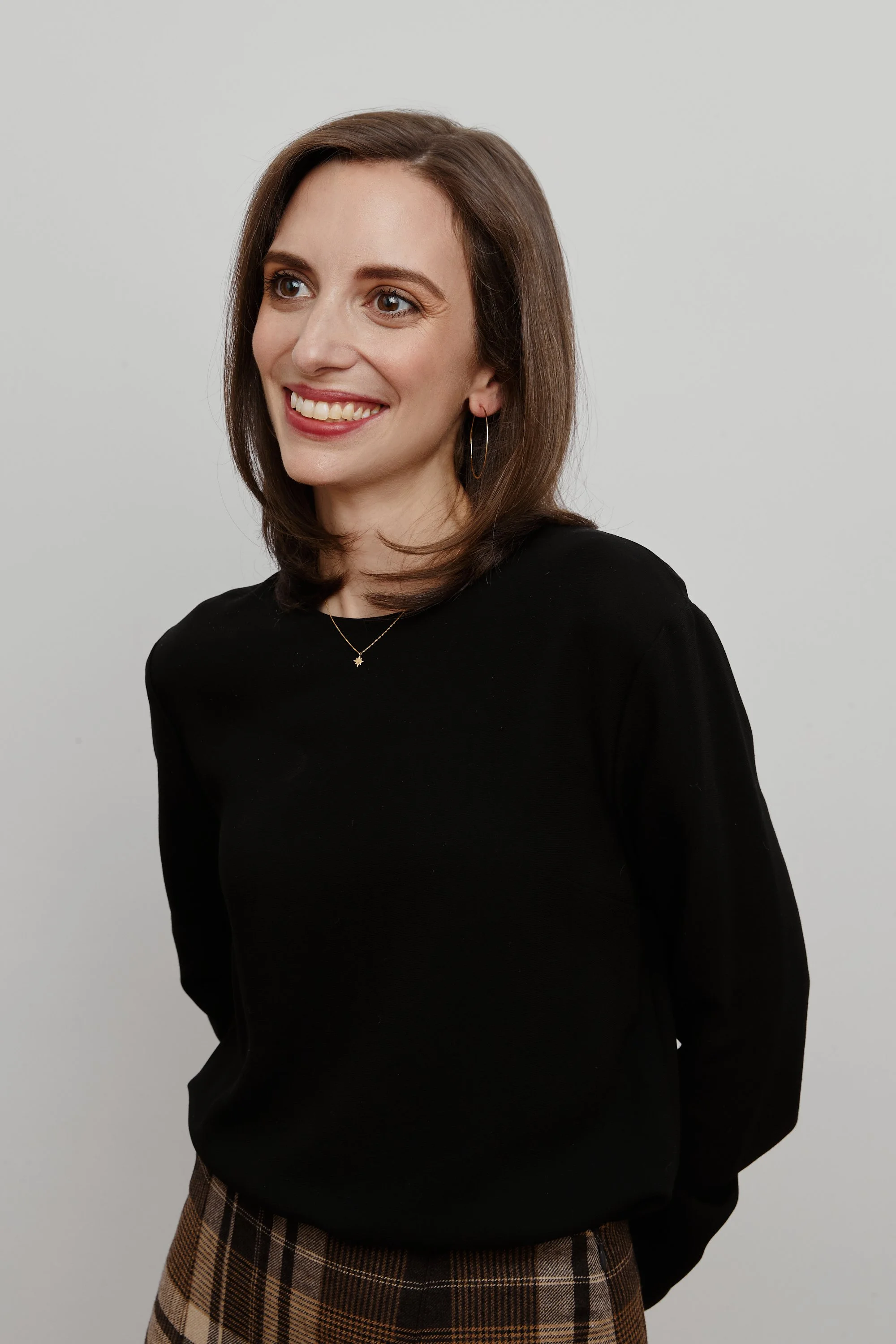 A woman with shoulder-length brown hair, smiling, wearing a black top, plaid skirt, and jewelry, standing against a plain light gray background.