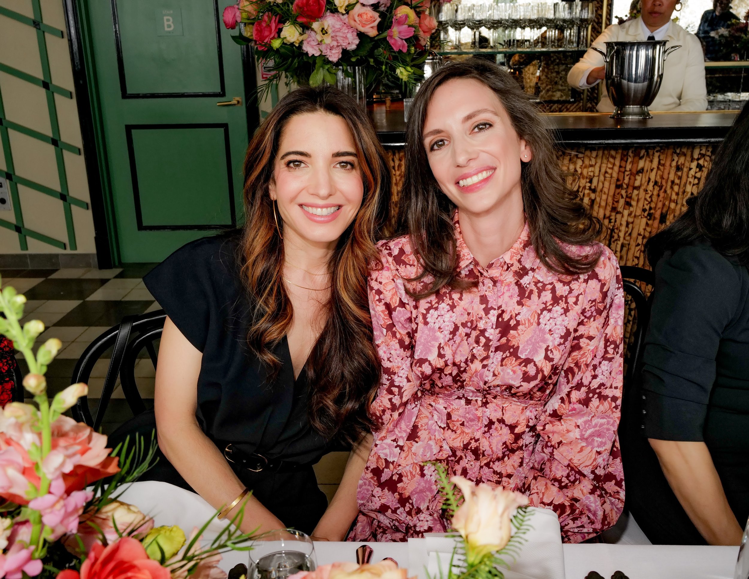 Two women smiling and sitting at a table during a celebration or gathering, with flowers and a decorated table in front of them.