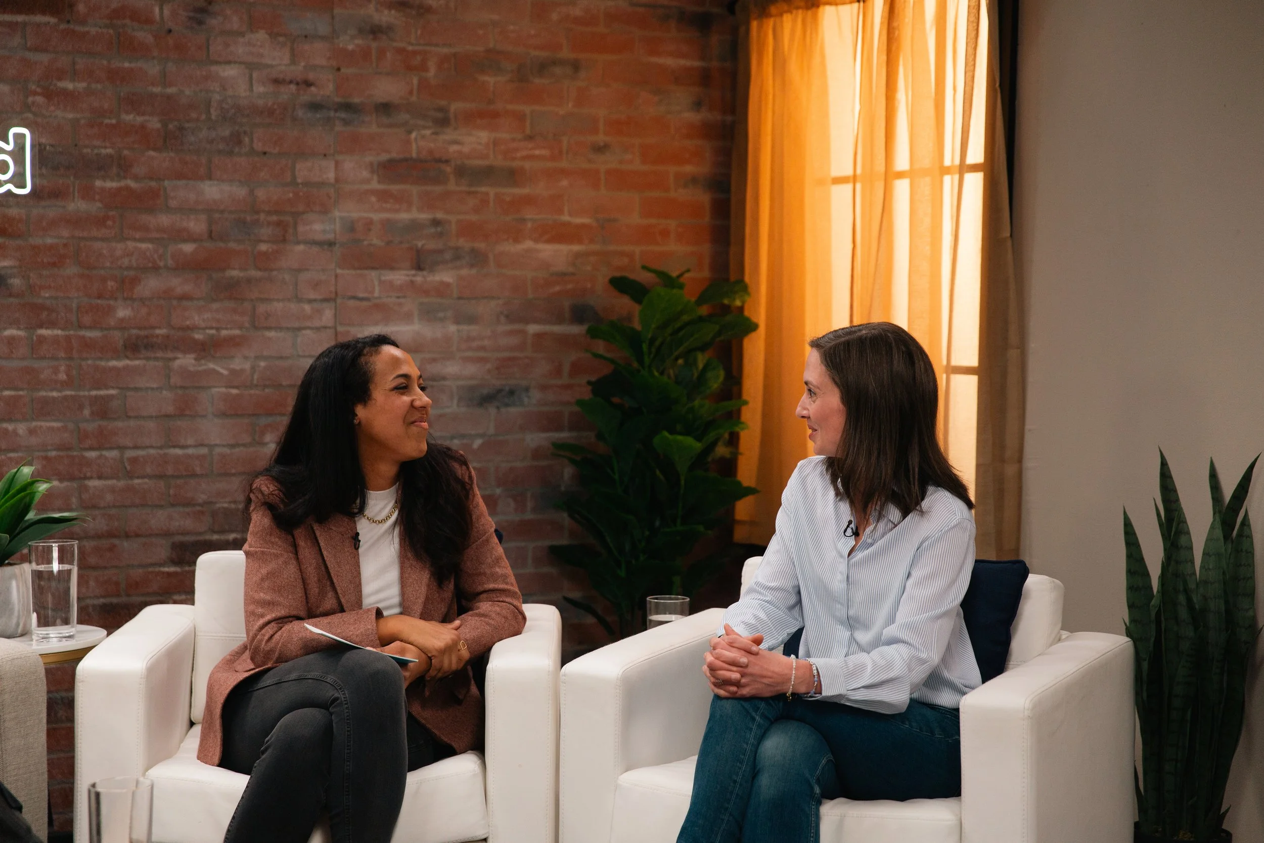 Two women sitting on white chairs having a conversation in a cozy room with a brick wall, green plants, and an orange curtain.