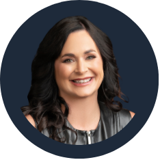 A smiling woman with dark wavy hair, wearing a sleeveless top and a leather jacket against a dark background