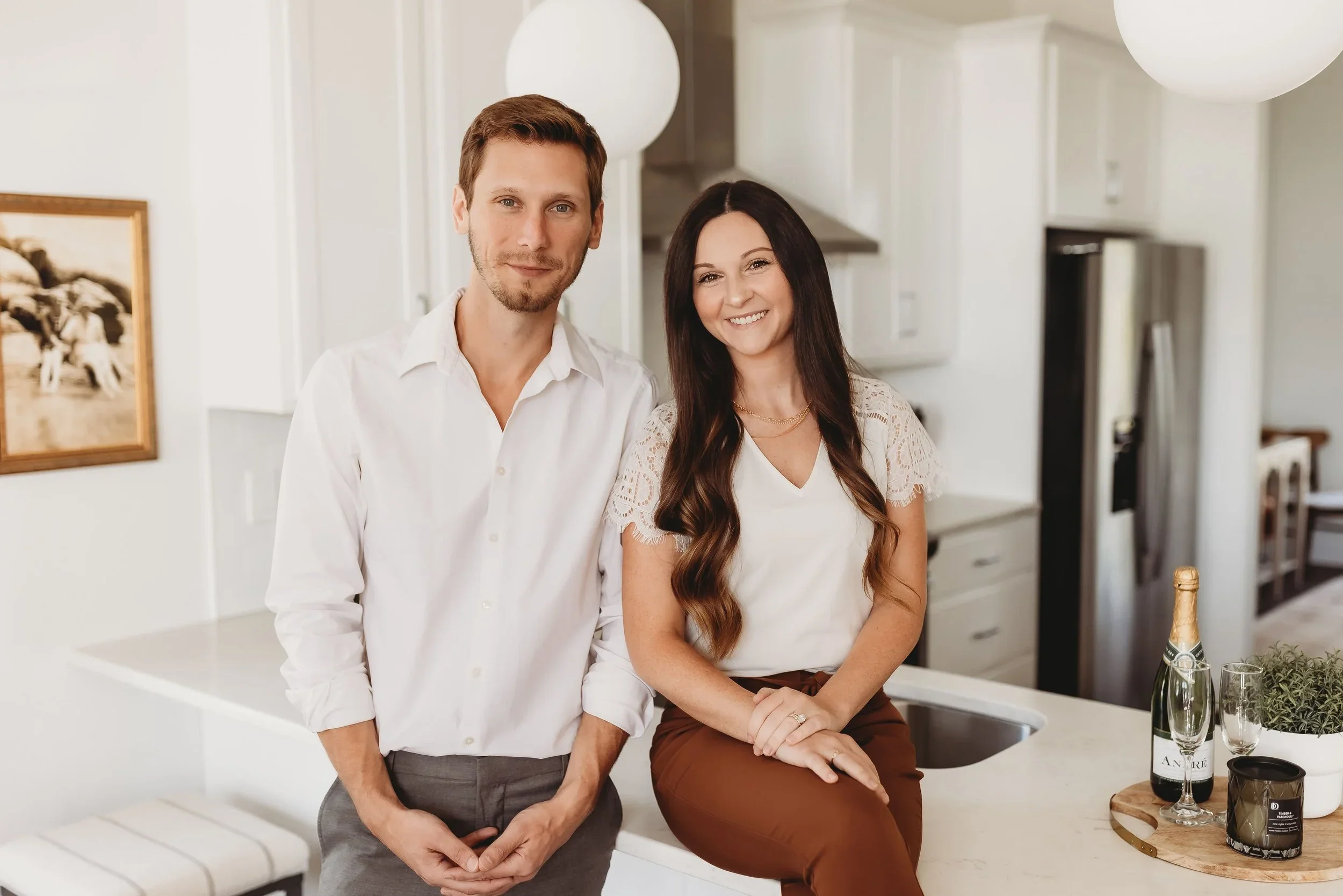 A smiling man and woman in a modern kitchen, with the woman sitting on the countertop near a bottle of champagne and glasses.