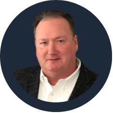 Headshot of a middle-aged man with short brown hair, wearing a dark blazer and white shirt, against a dark background.