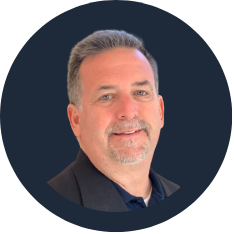 Portrait of a middle-aged man with short gray hair and a beard, wearing a dark business suit, against a dark geometric background.
