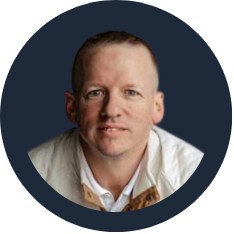 A portrait of a man with short light hair, wearing a white shirt and a beige jacket, against a dark background.