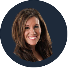 A woman with long brown hair smiling, wearing a dark top, against a dark background.