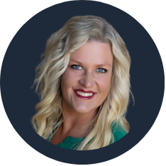 Portrait of a smiling woman with blonde, wavy hair, wearing a dark top and a teal shirt, against a dark background.