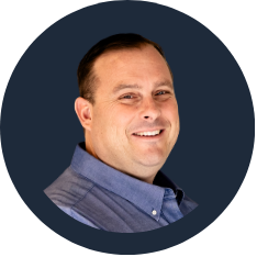 A man with short dark hair smiling, wearing a blue button-up shirt, against a dark background.