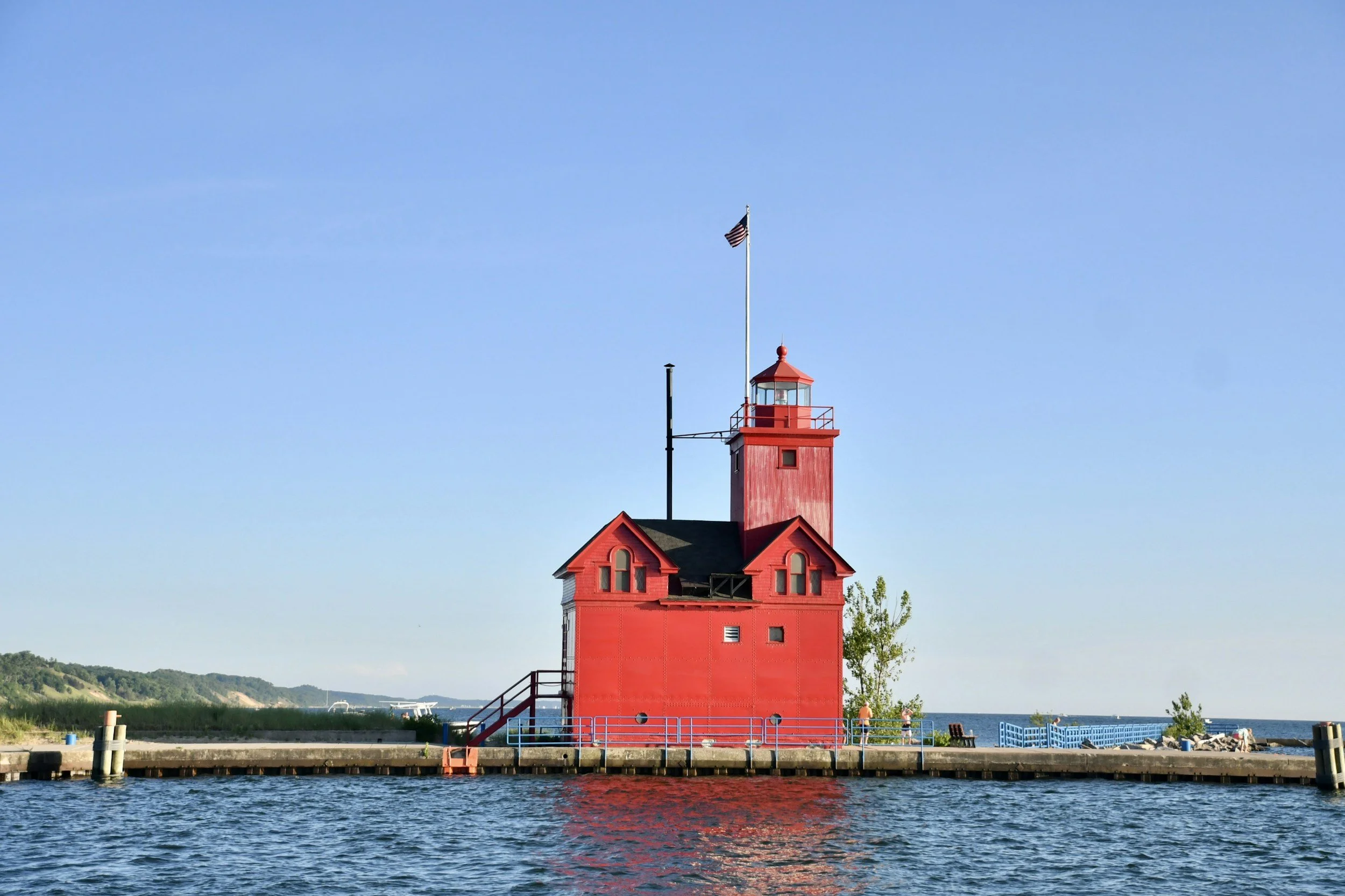 A red lighthouse on a pier with water in the foreground, blue sky, and some greenery in the background.