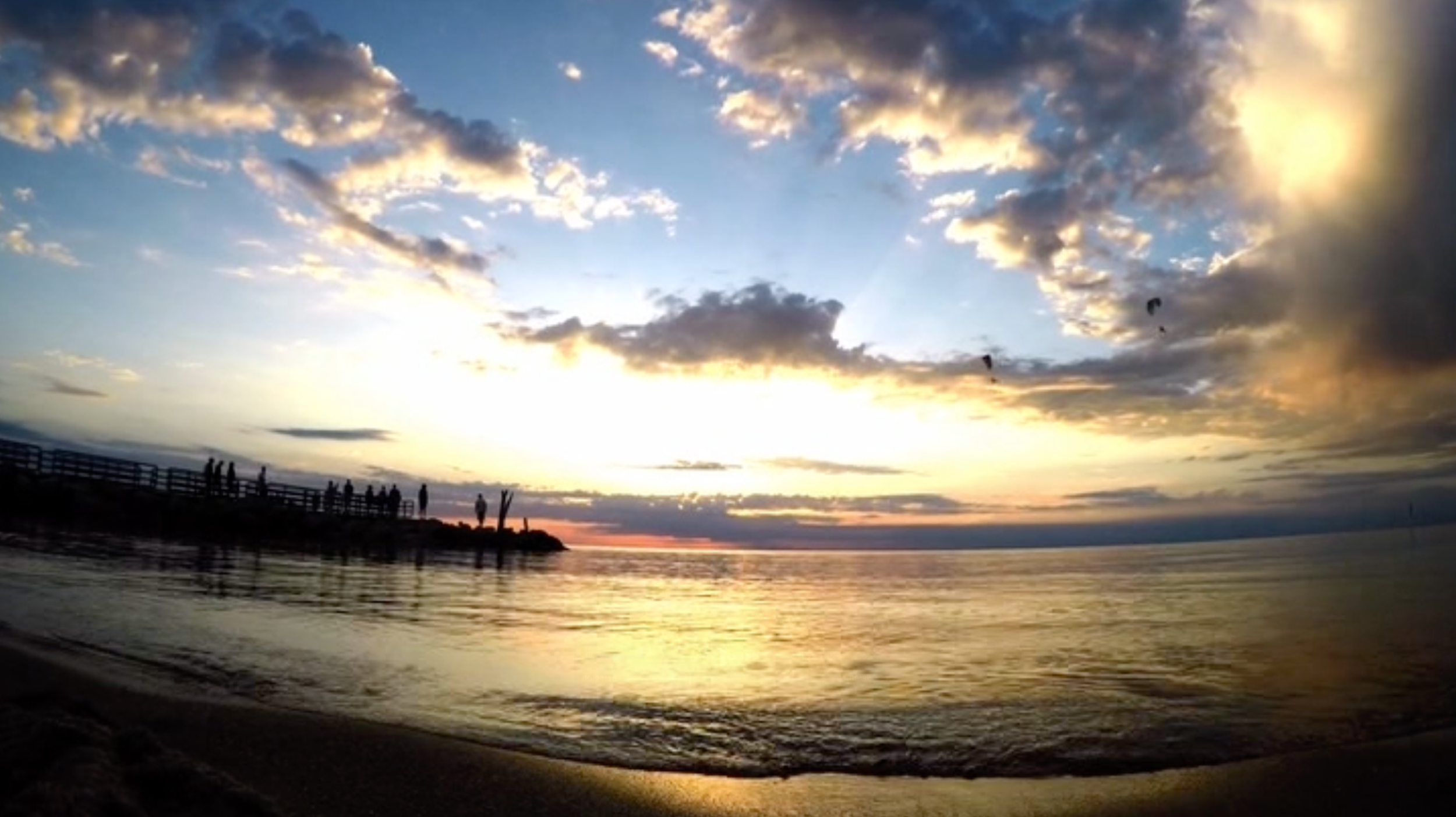 Sunset at the beach with silhouetted people on a pier, clouds in the sky, and calm water reflecting the colors of the sunset.