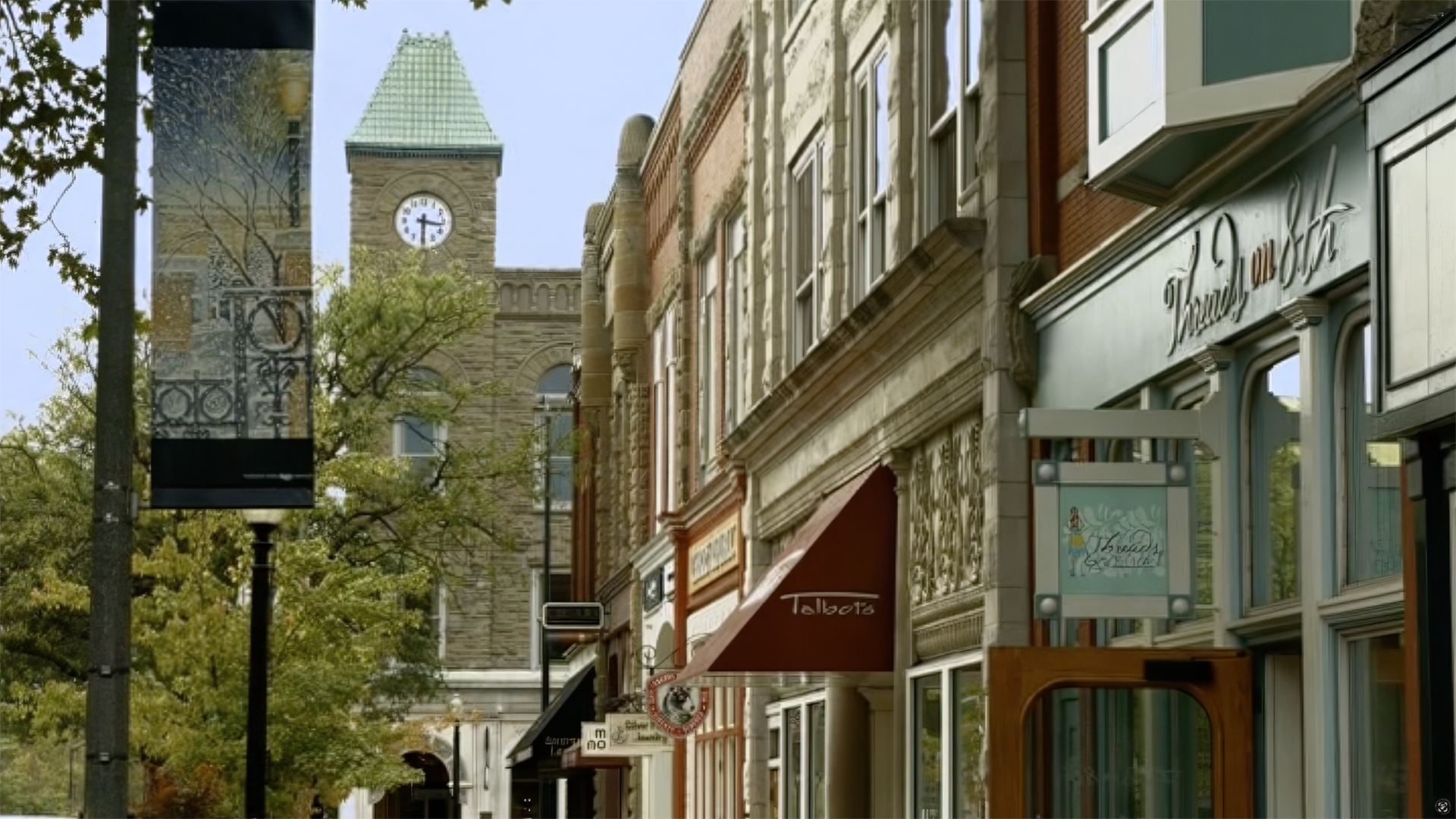 Street view of historic downtown with storefronts, a clock tower in the background, and trees lining the sidewalk.