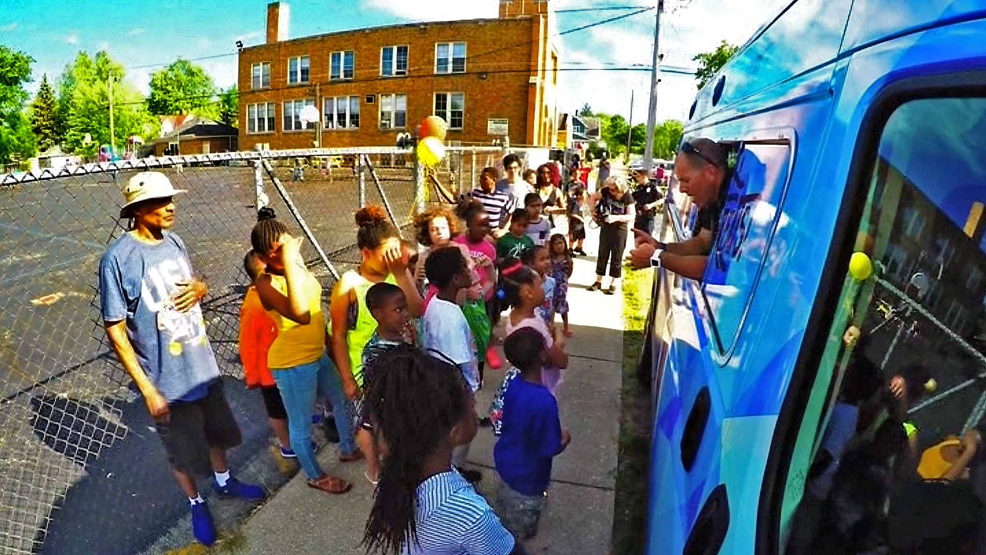 A group of children and adults gathered outside near a blue bus, listening to a man speaking from the bus window, on a sunny day with a building and trees in the background.