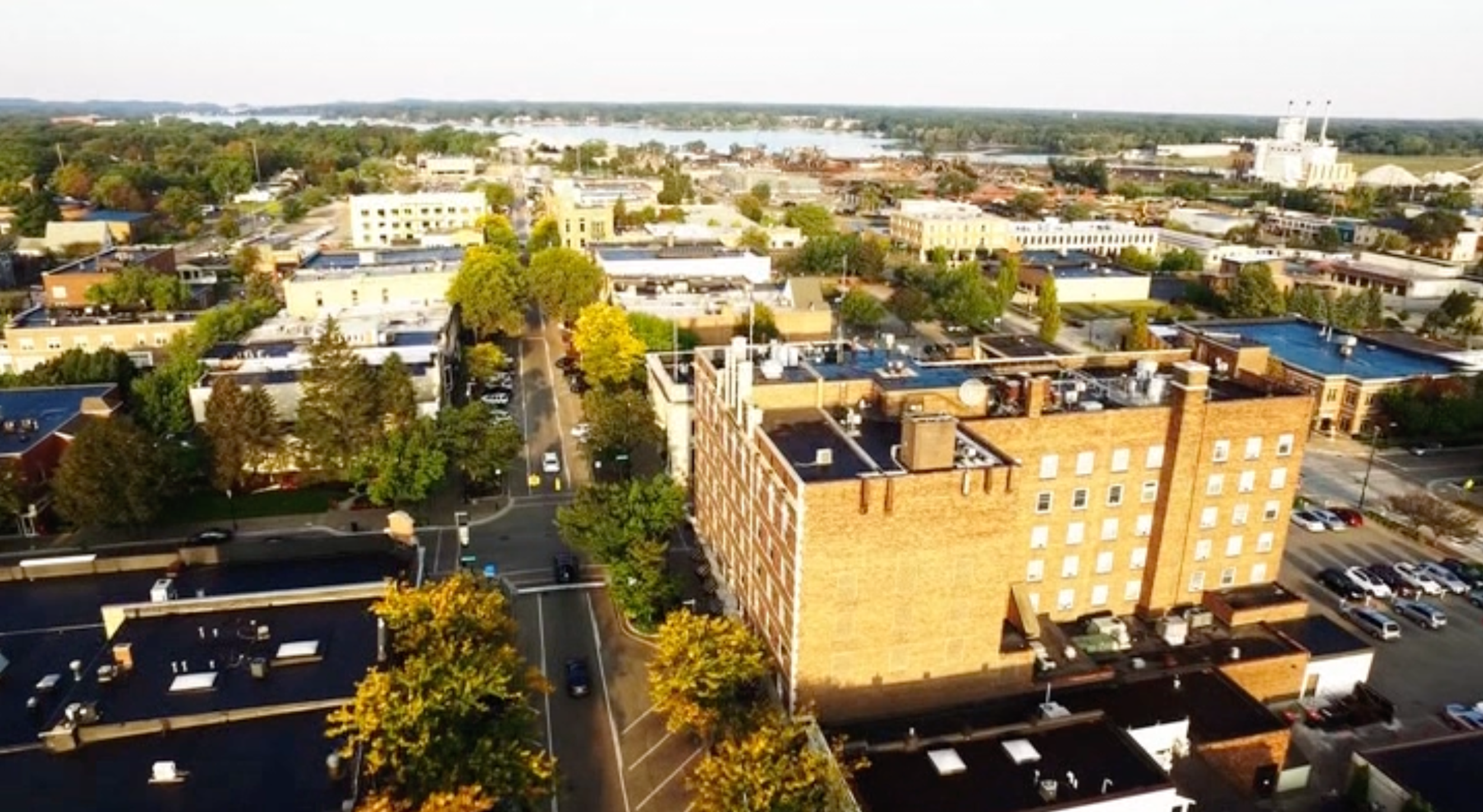 Aerial view of a cityscape with buildings, streets, parked cars, trees, and a large body of water in the background.