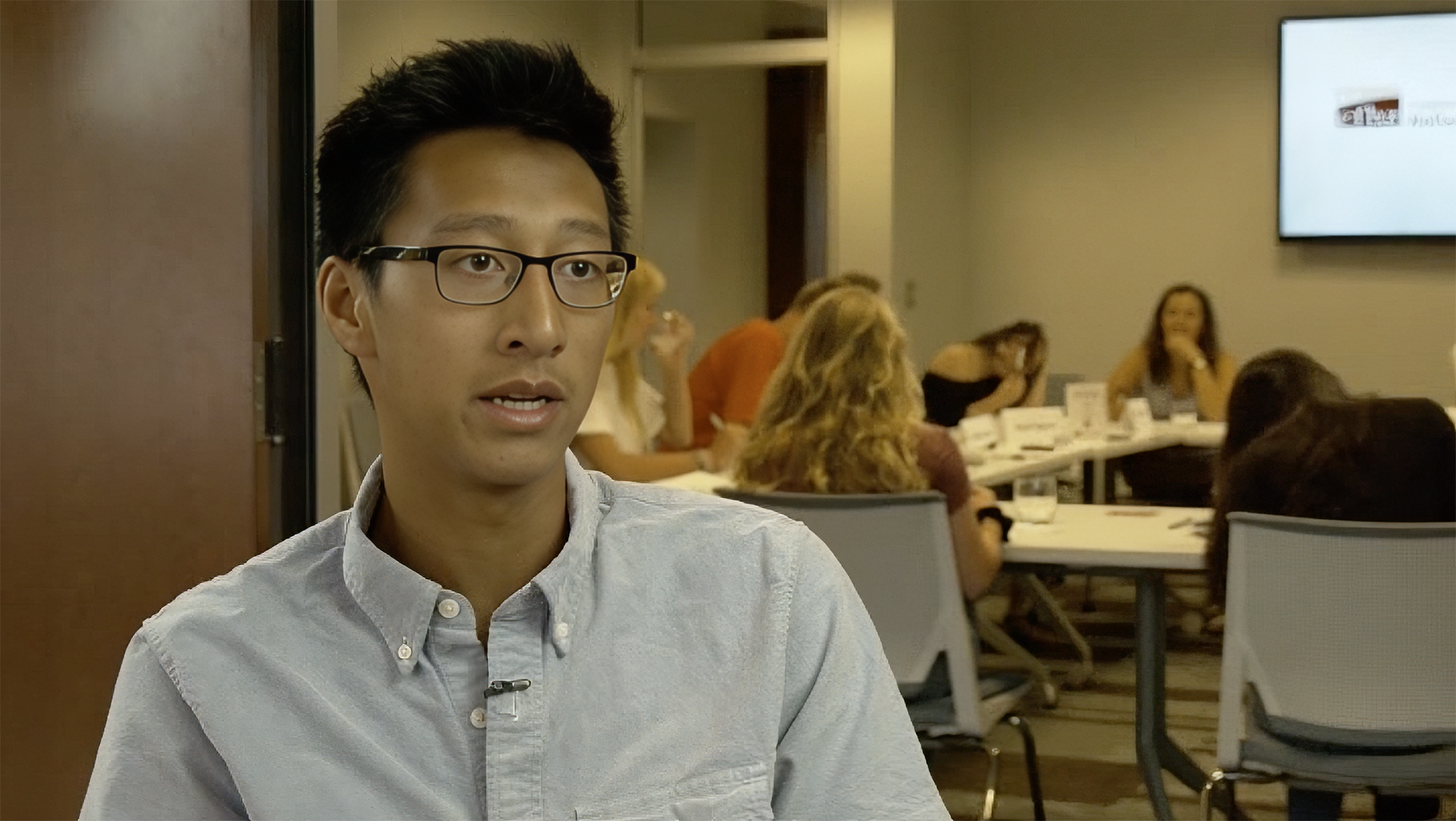 A young man with glasses and a white shirt being interviewed in a conference room, with a group of people sitting at a long table in the background.