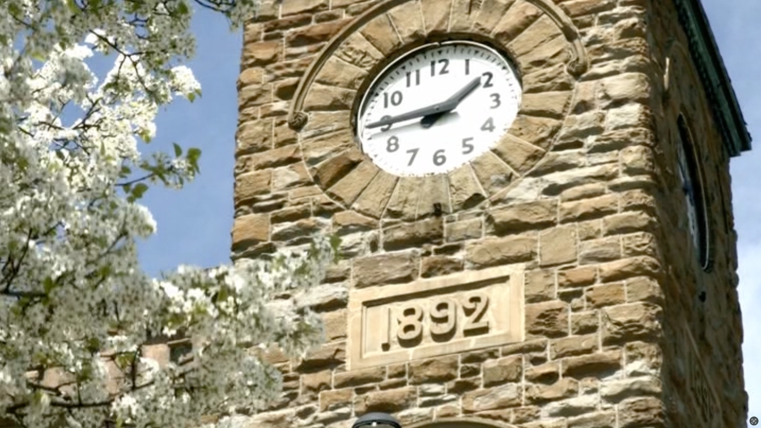A clock tower made of brown brick with a white clock face showing the time as 1:48, and the year 1892 inscribed on a stone plaque below the clock. White flowering tree branches are visible in the foreground.