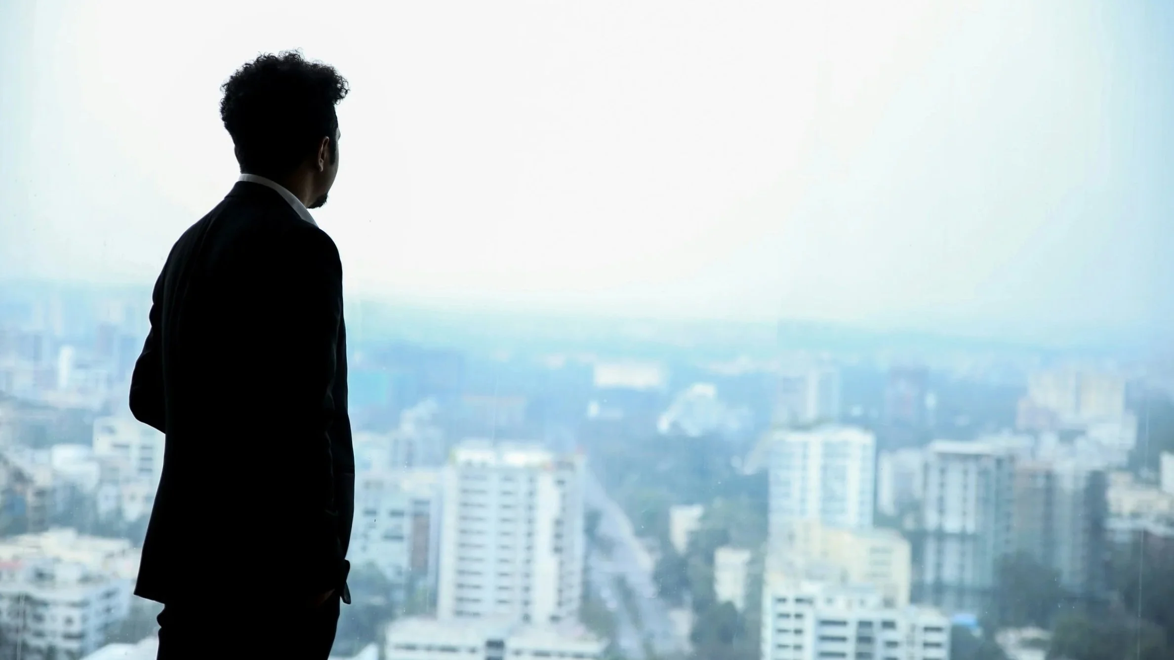 A man in a suit standing indoors, looking out a large window at a cityscape with tall buildings.