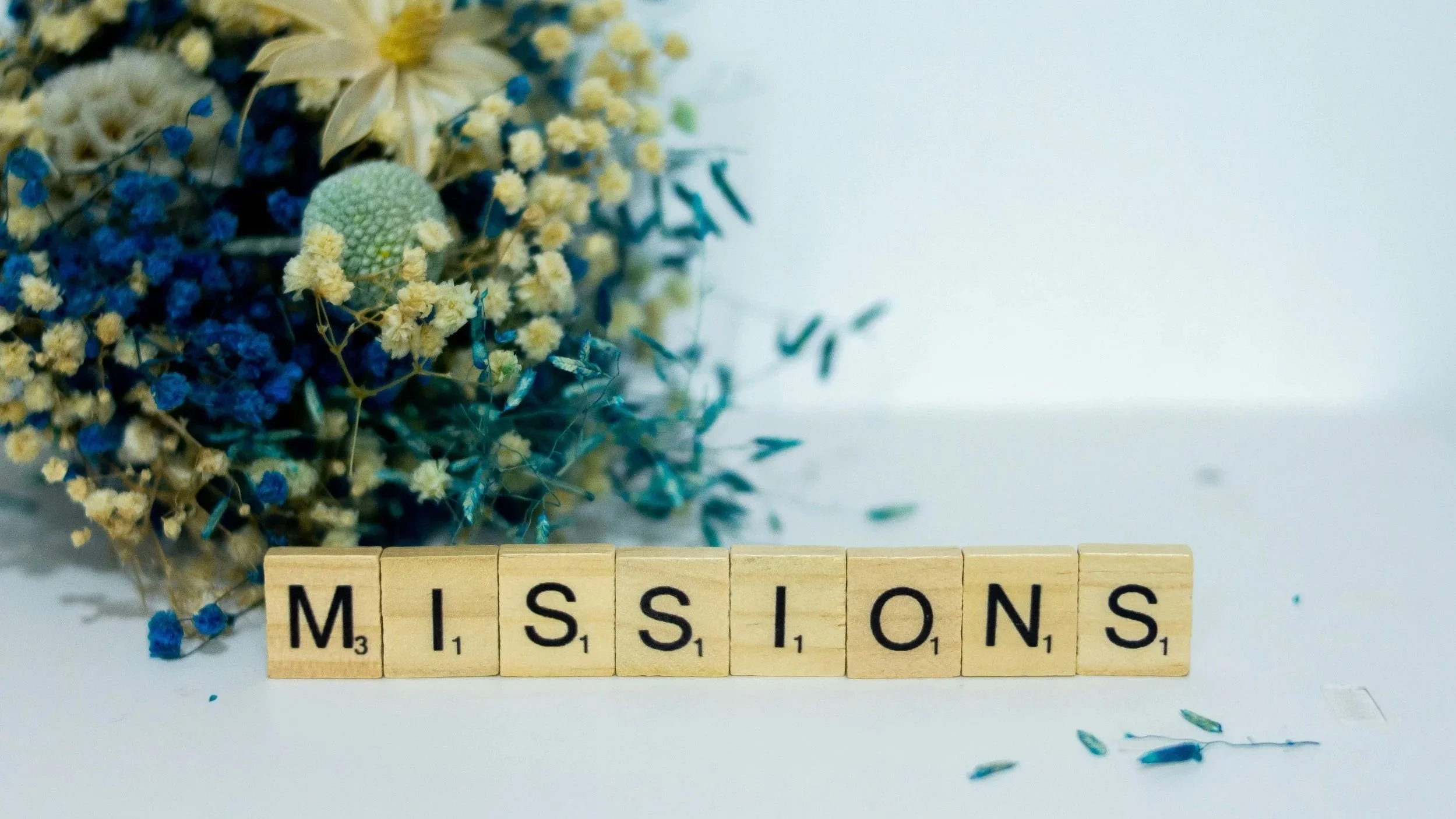 Scrabble tiles spelling 'MISSIONS' with a bouquet of yellow and blue flowers in the background.