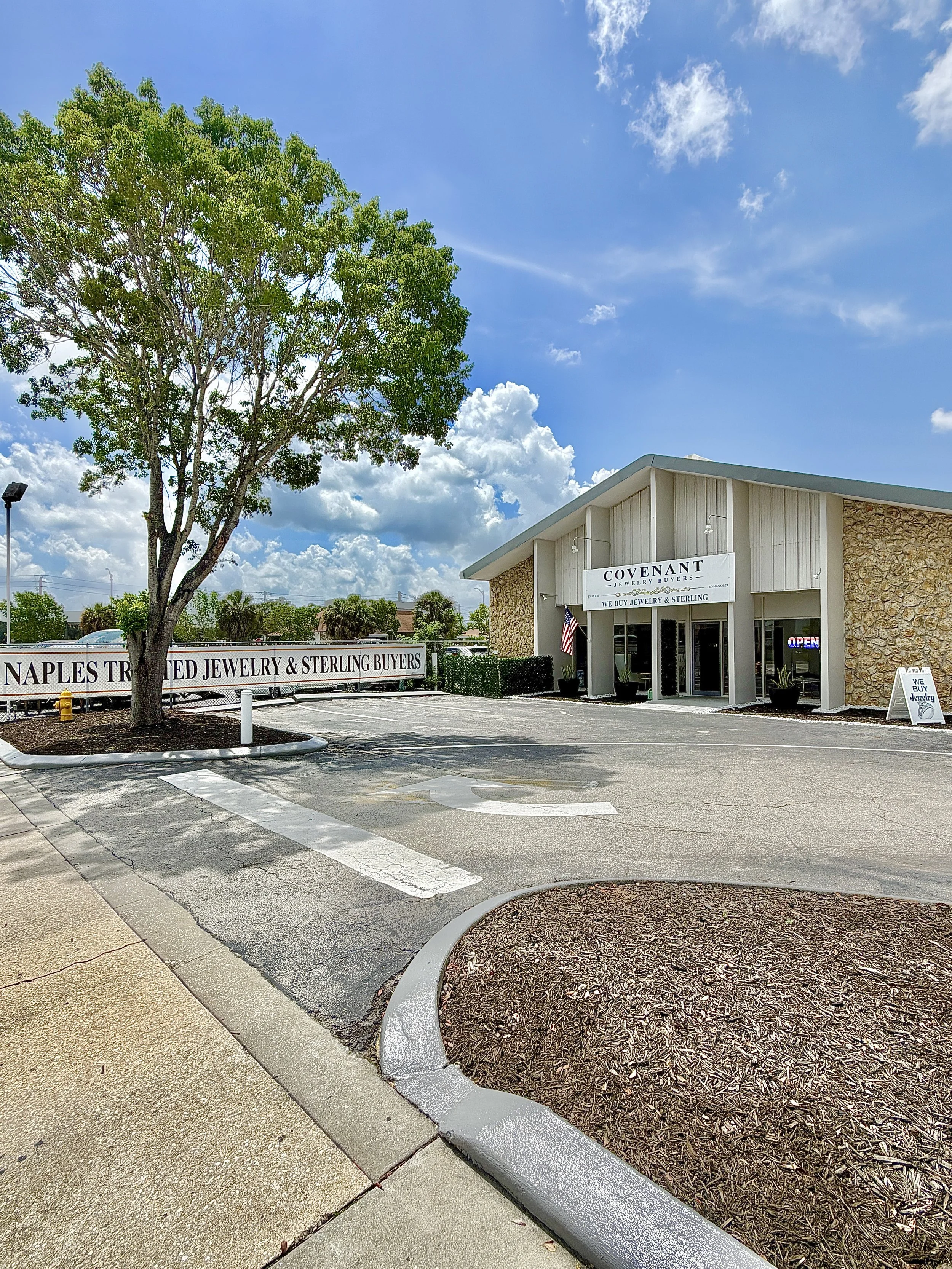 The image shows the exterior of a jewelry store called 'Covenant Jewelry Buyers' with a sign that states they buy jewelry and sterling silver. There is a parking lot with a curb, a small landscaped area with mulch, and a large tree on the left side. The sky is partly cloudy with blue sky and white clouds. An 'Open' sign is visible in the store window.