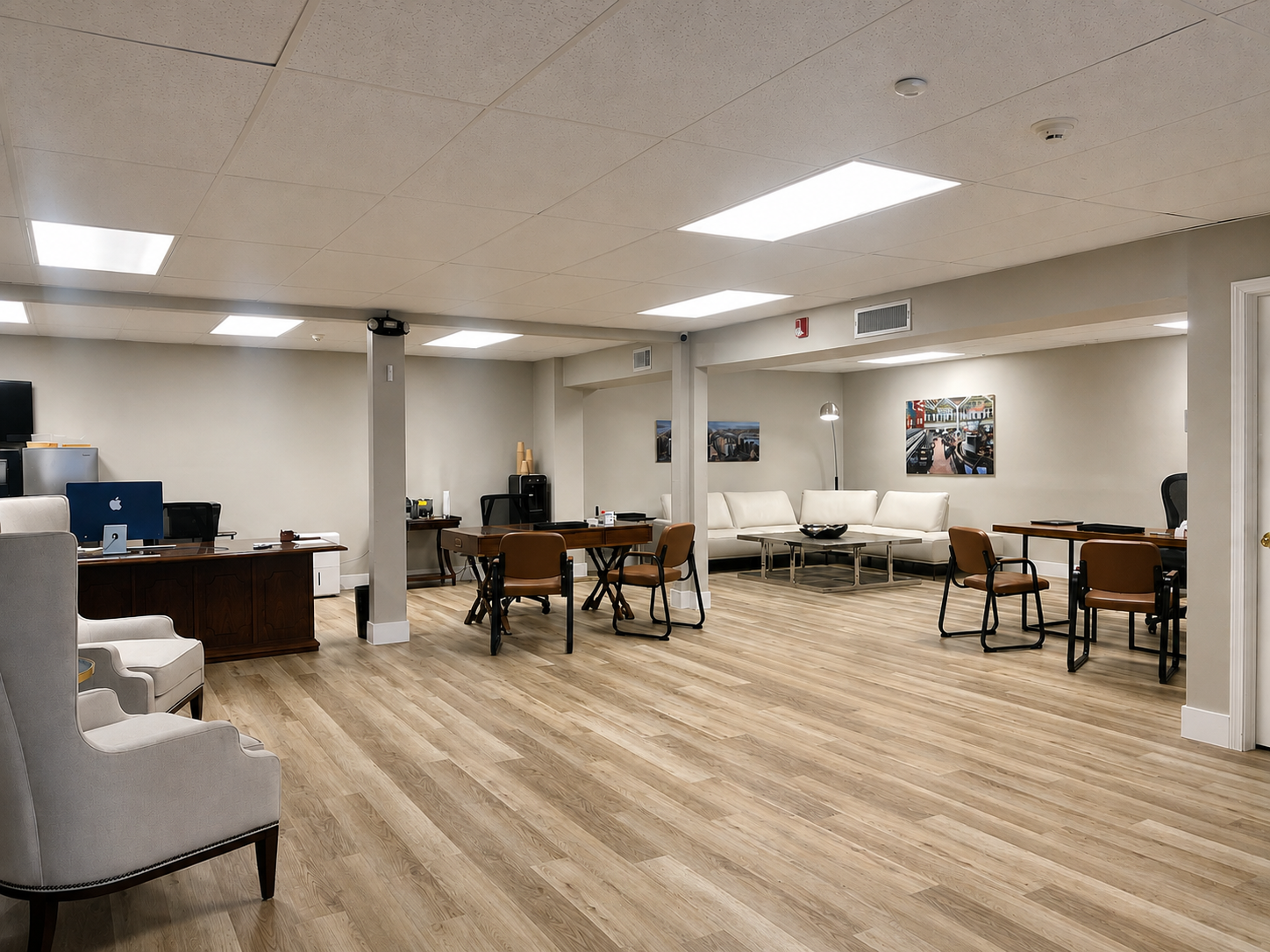 Open office lounge area with wooden flooring, white armchairs, desks, and a white sectional sofa with coffee tables. Wall decor includes framed pictures, and overhead lighting is visible.