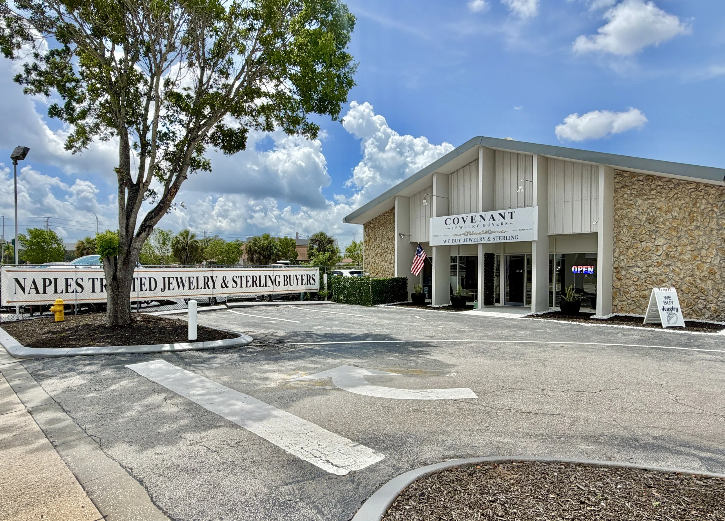 The image shows a jewelry store named Covenant Jewelry with a sign that says they buy jewelry and sterling silver. The store has a light-colored facade with stone accent walls and a small American flag outside. The parking lot in front is mostly empty, with a tree and some bushes nearby, under a blue sky with scattered clouds.