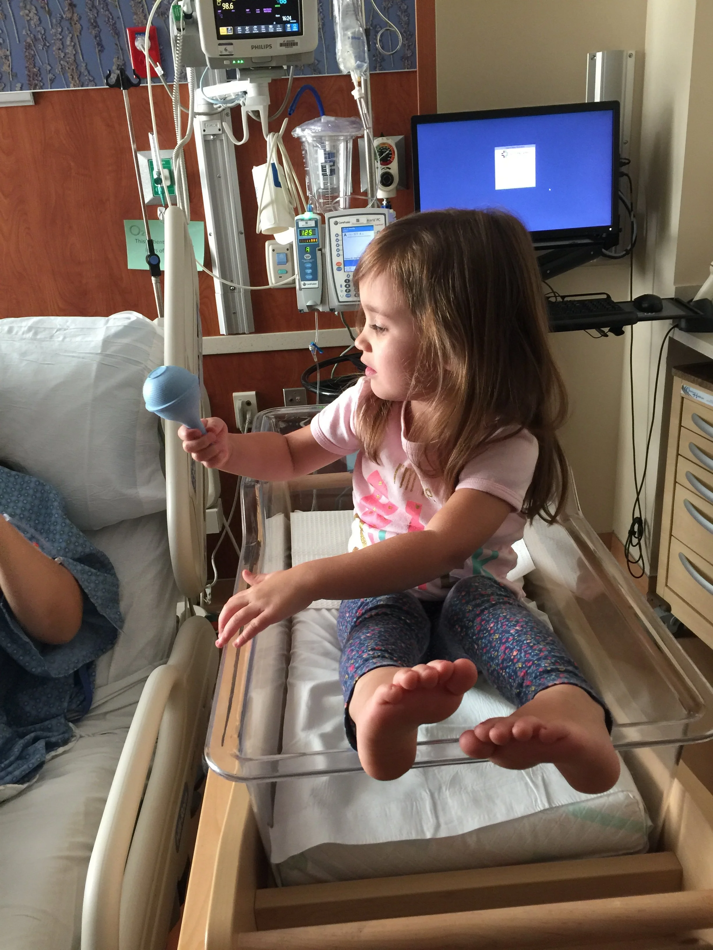 A young girl sitting on a hospital bed for a newborn with her legs and feet extending over a metal tray. Medical equipment, monitors, and a computer are visible in the room.