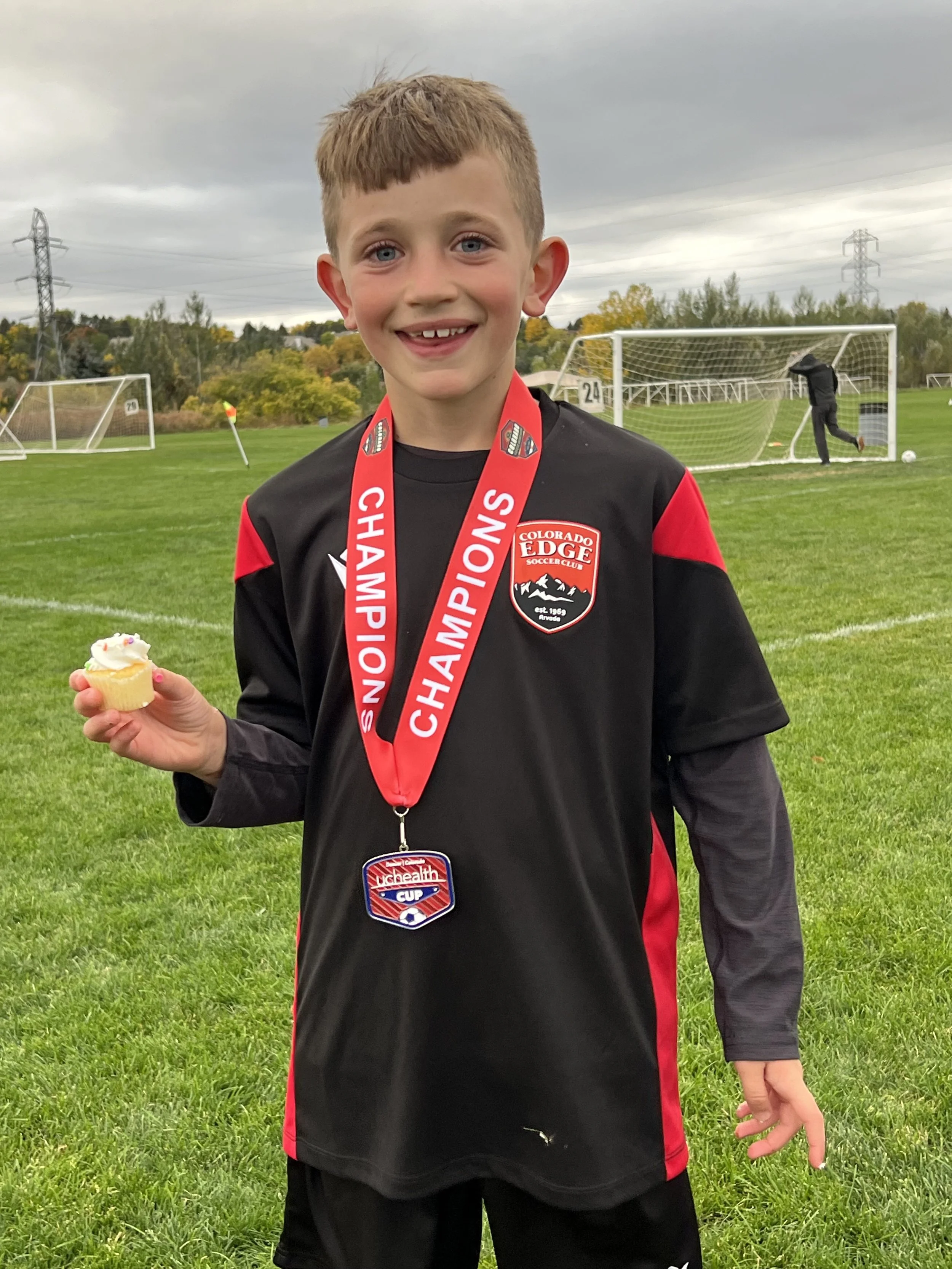 A young boy with short, light brown hair and blue eyes smiling, wearing a black and red soccer jersey with a Colorado Edge Soccer Club badge, holding a cupcake with white frosting and colorful sprinkles, on a soccer field with goalposts and cloudy sky in the background.