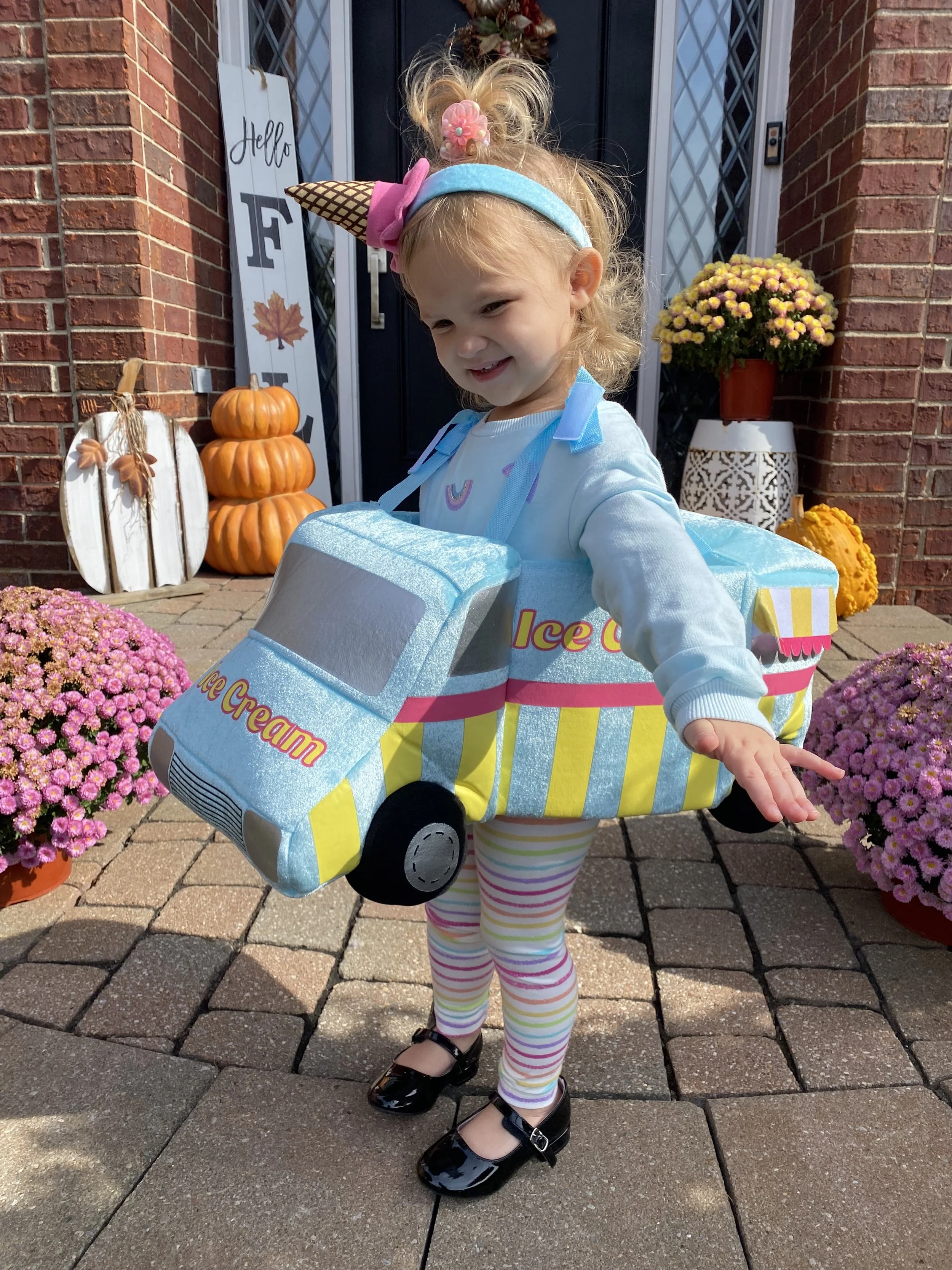 Young girl dressed as an ice cream truck, smiling outdoors with fall decorations and flowers in the background.