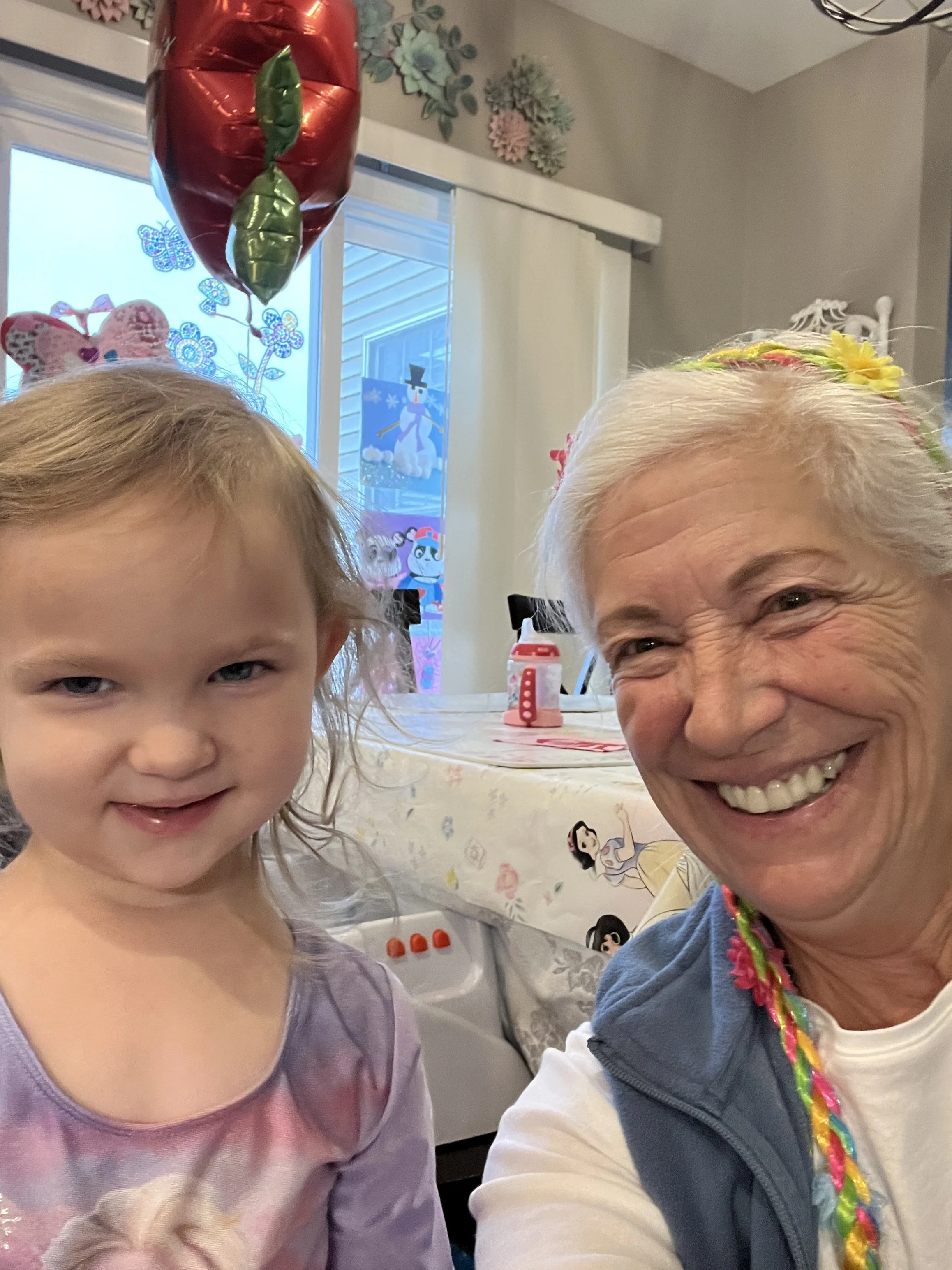 A young girl with blond hair and a woman with gray hair smiling for a selfie at a birthday celebration. In the background, there is a table with a Disney princess themed tablecloth, birthday balloons, and holiday decorations including a snowman windo