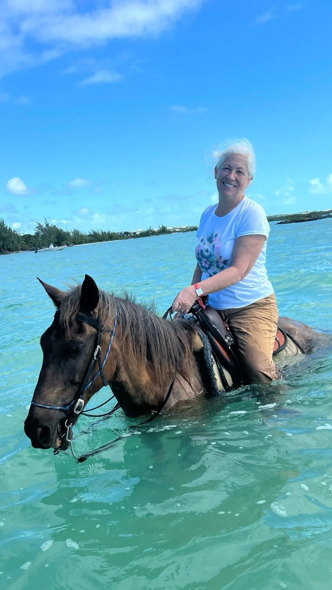 A woman with white hair riding a horse through clear, shallow water on a sunny day, with a blue sky and green shoreline in the background.