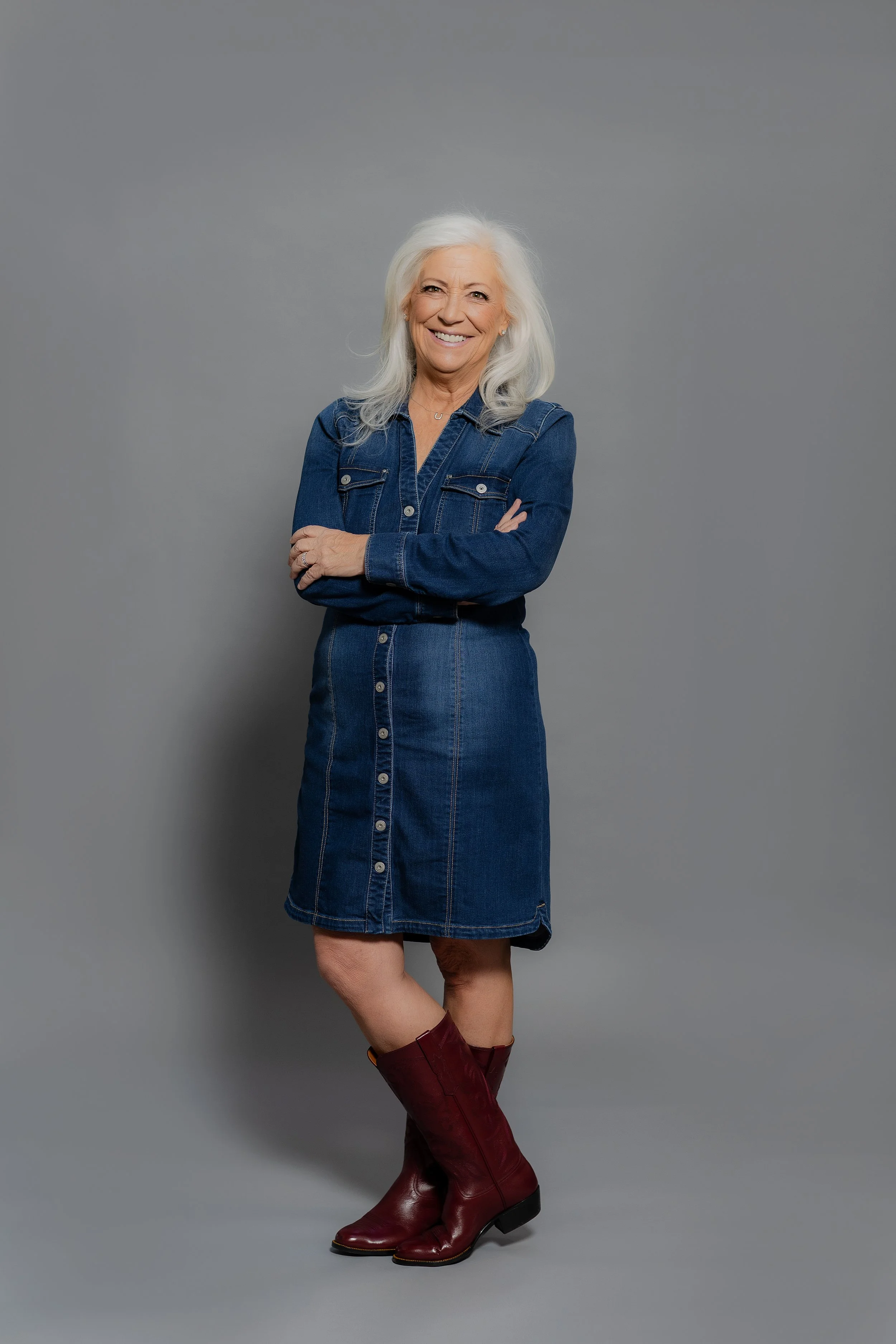 A woman with long white hair is smiling and standing with her arms crossed against a plain gray background. She is wearing a denim dress and maroon knee-high boots.