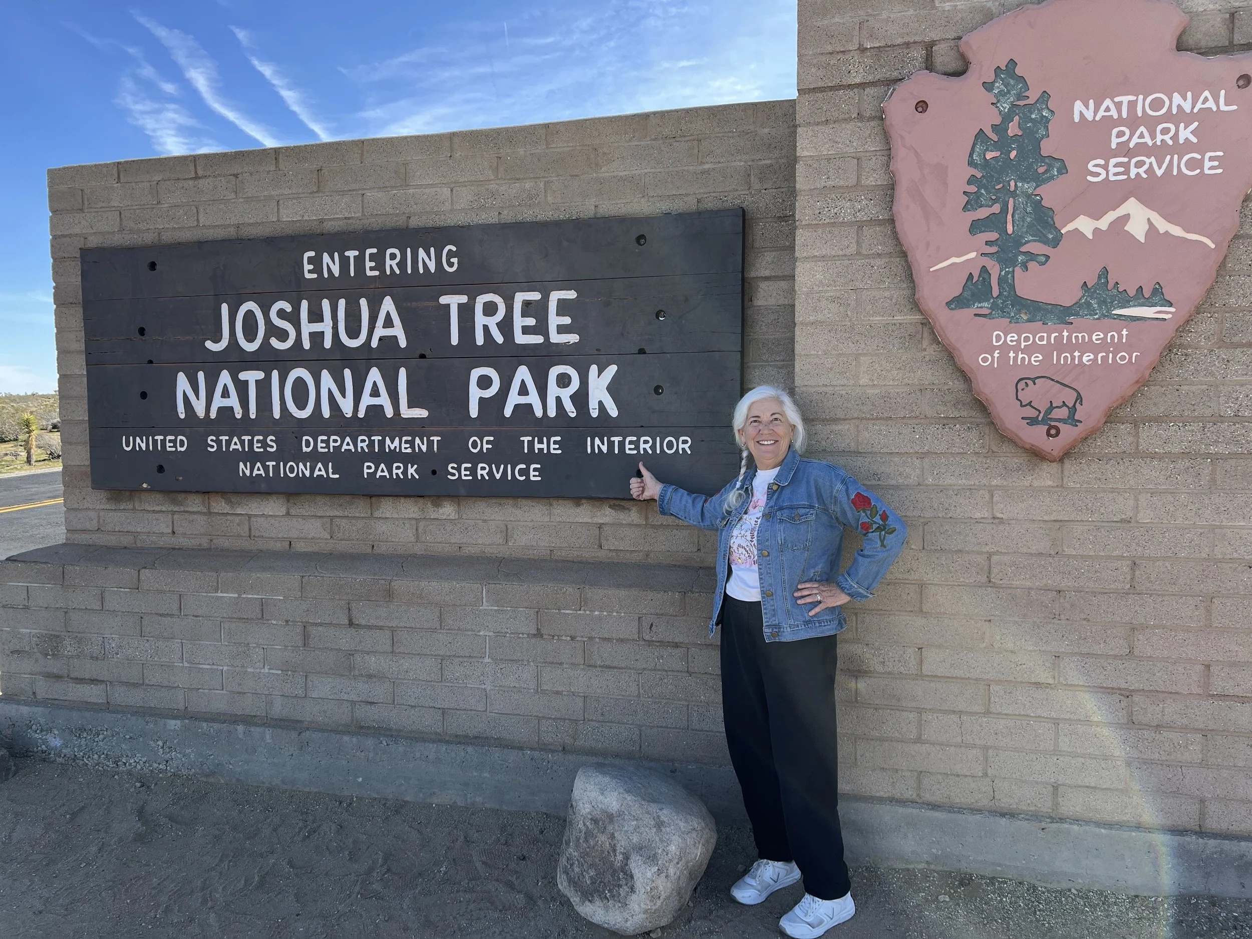 A smiling woman with white hair in a denim jacket standing next to the entrance sign for Joshua Tree National Park, with a map and park service emblem on a brick wall background.