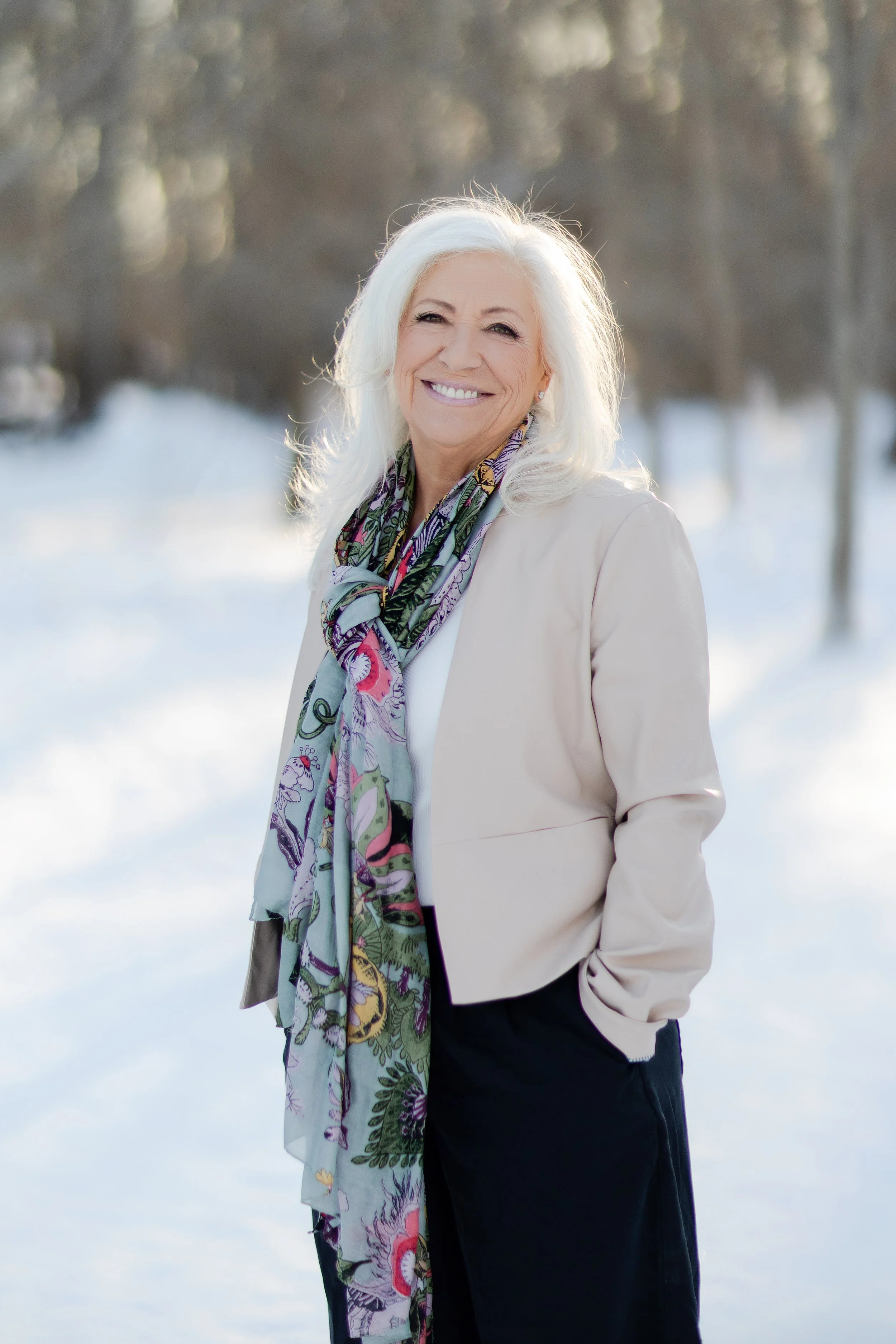 A mature woman with long white hair smiling outdoors in a snowy landscape, wearing a light-colored blazer, a colorful scarf, and dark pants.