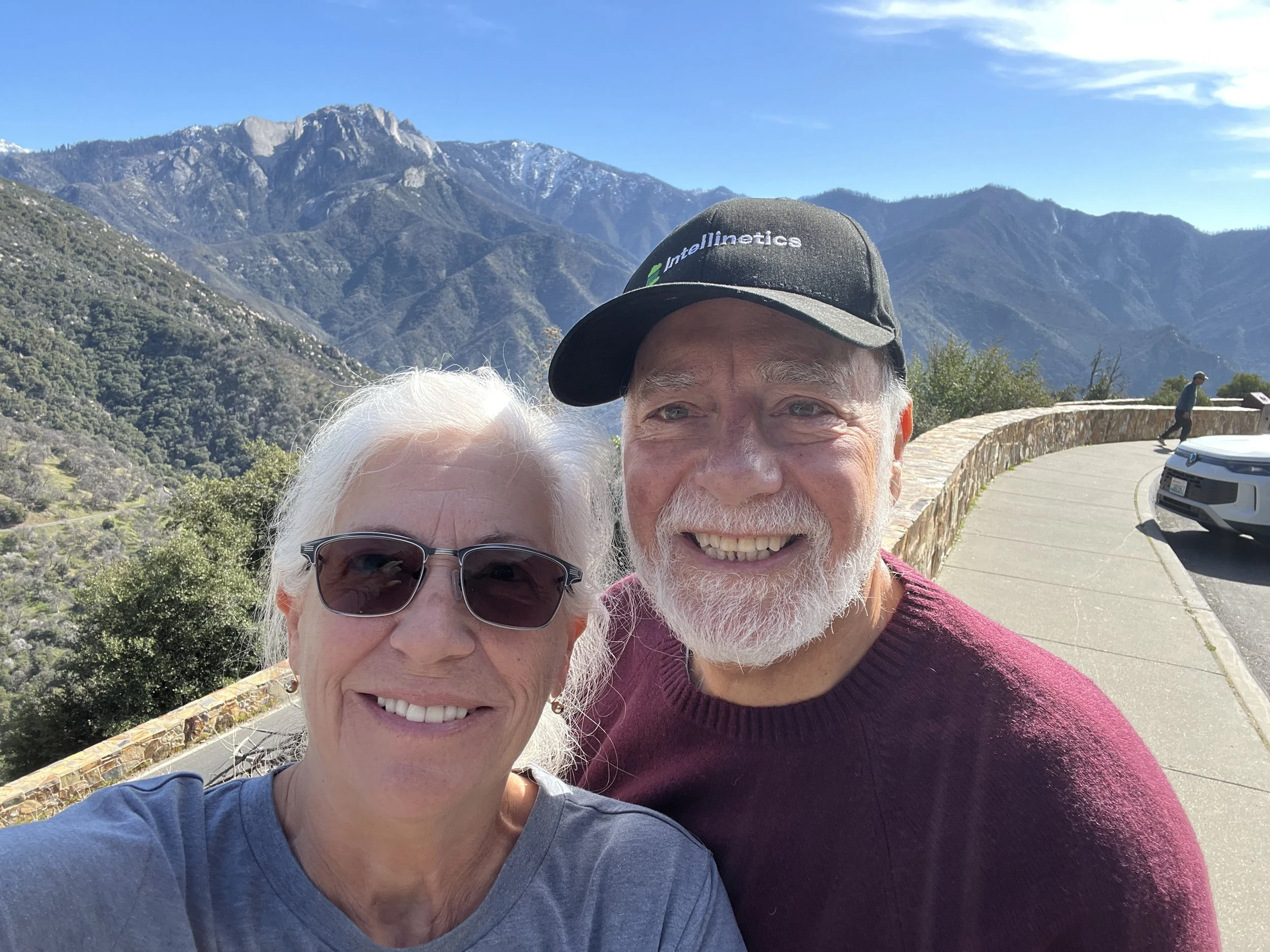 Close-up of a woman with white hair and sunglasses and a smiling man with a beard and black cap, in front of a mountainous landscape with clear blue sky.