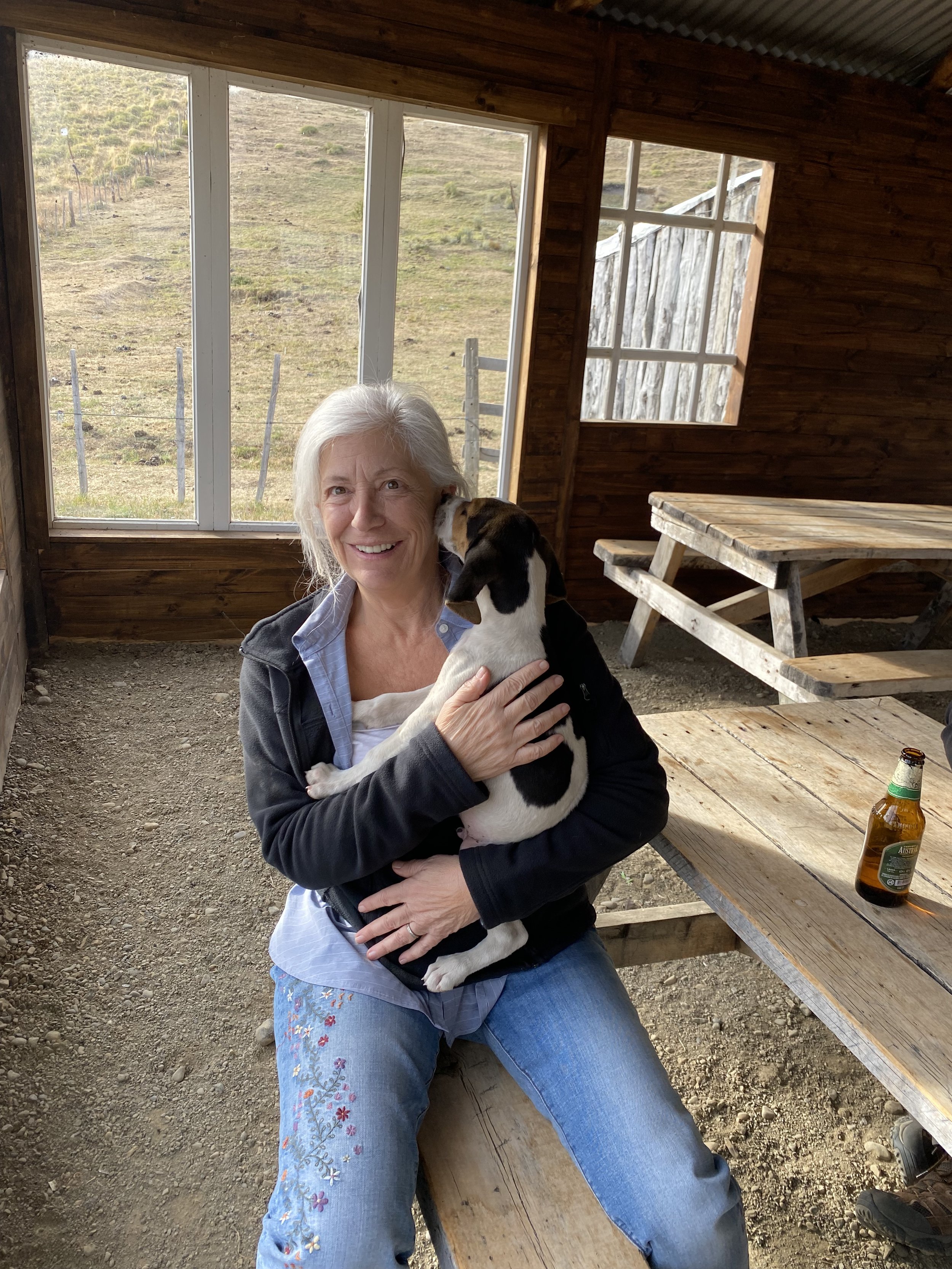 An older woman with white hair, wearing a black jacket and light blue denim jeans with floral embroidery, is sitting and holding a black and white puppy. The woman is smiling, and the puppy is nuzzling her face. They are inside a rustic wooden struct