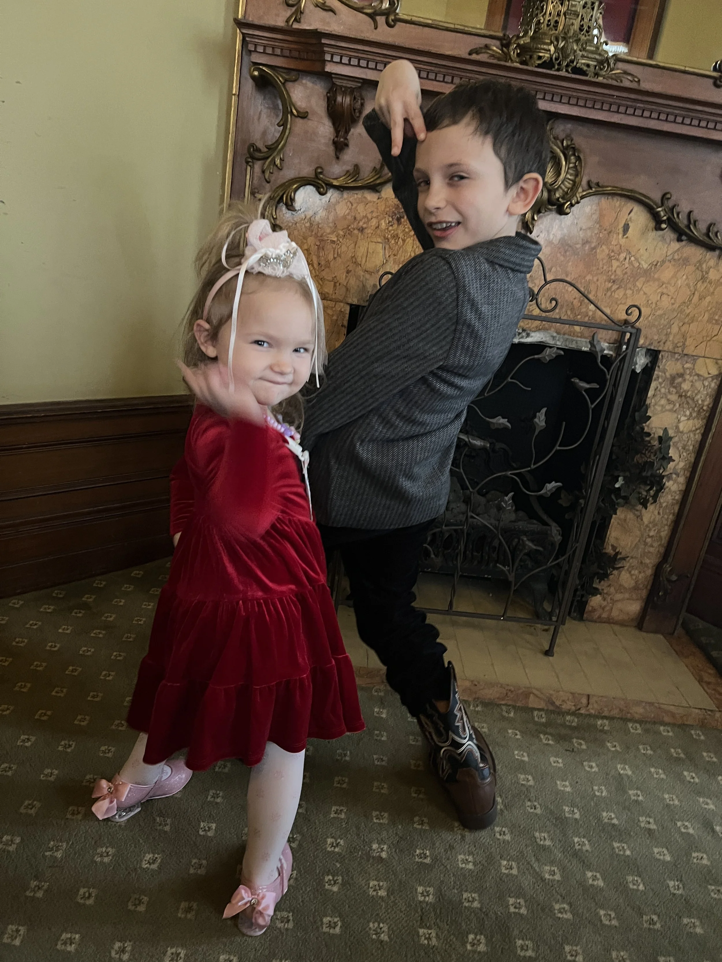 A young girl and boy posing indoors, with the girl in a red velvet dress, white tights, pink ballet shoes, and a decorative headband, and the boy in a dark striped jacket, black pants, and cowboy boots. They are standing in front of a fireplace with 