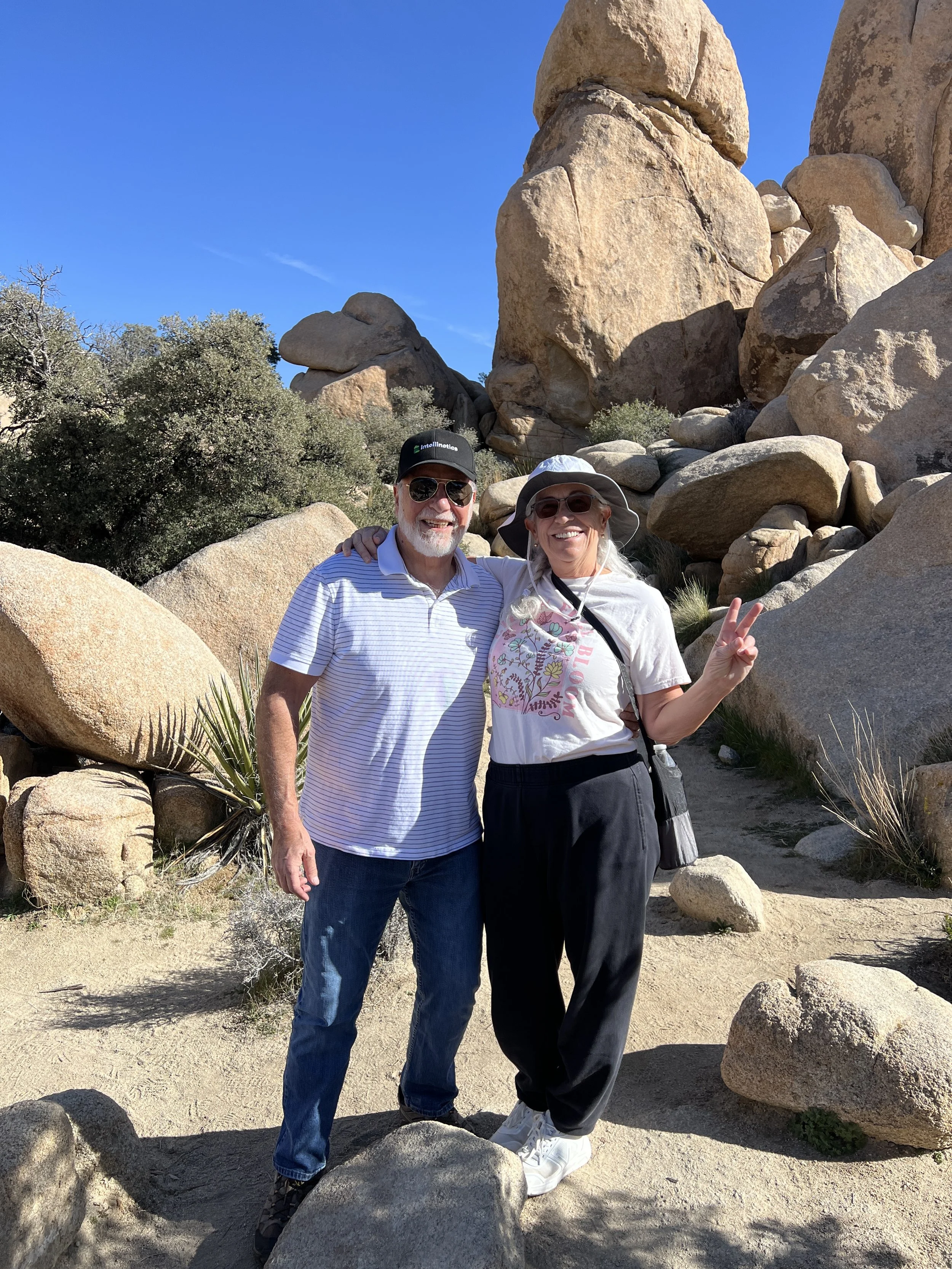 A smiling couple standing in front of large rocks and desert vegetation under a clear blue sky. The man is wearing a striped polo shirt, glasses, a black cap, and jeans. The woman is wearing a white hat, sunglasses, a white shirt with colorful design