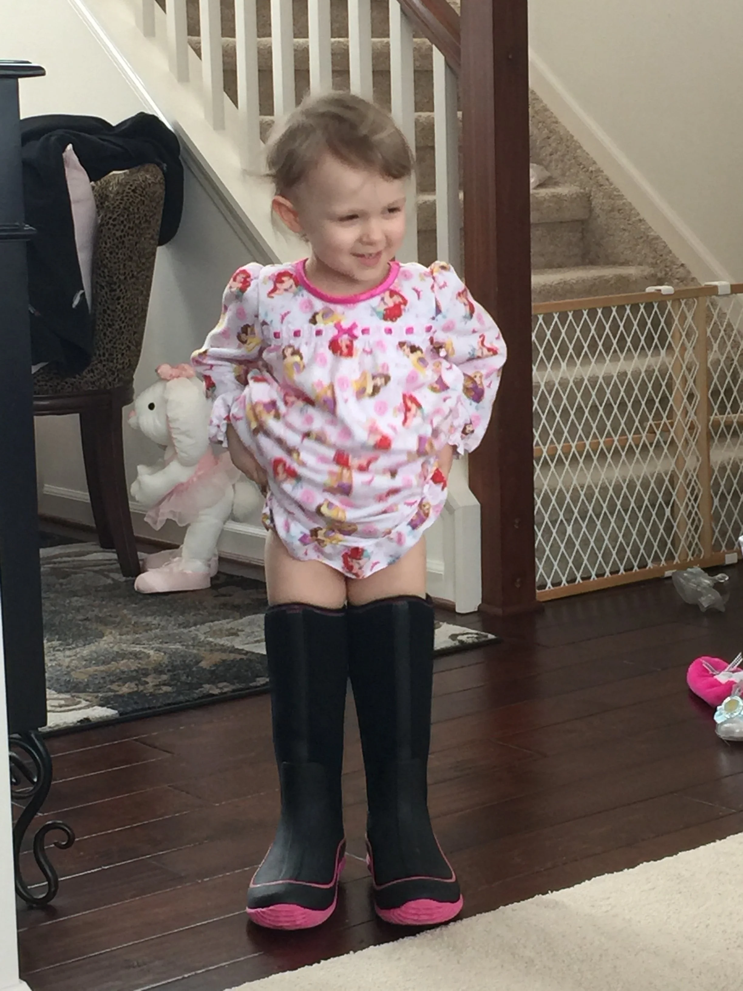 Kids standing in front of a mirror, girl with a patterned pajamas and black boots, smiling with a pair of plush slippers nearby.