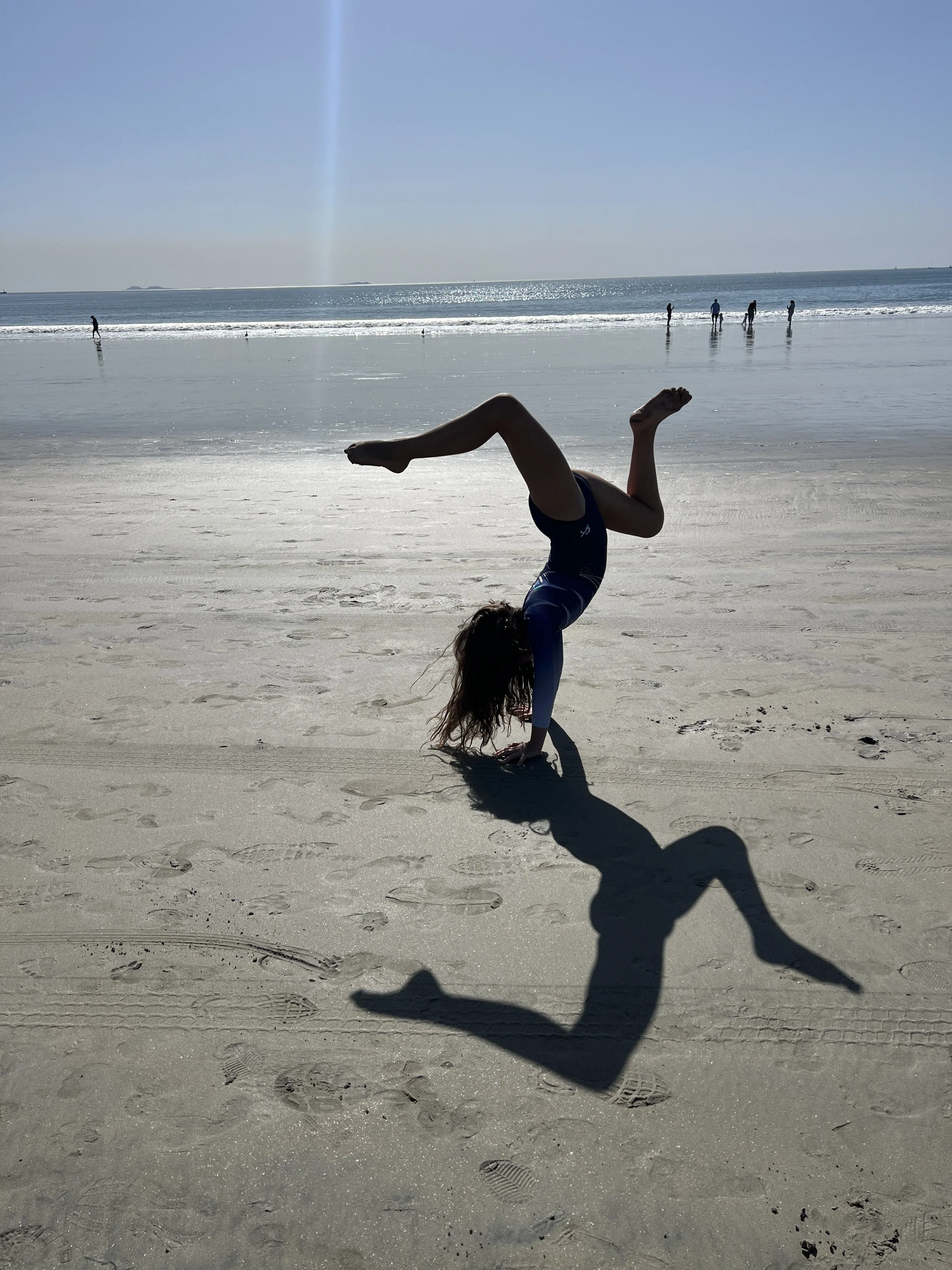 A person doing a handstand on the beach with the ocean and several people walking along the shoreline in the background, under a clear blue sky.