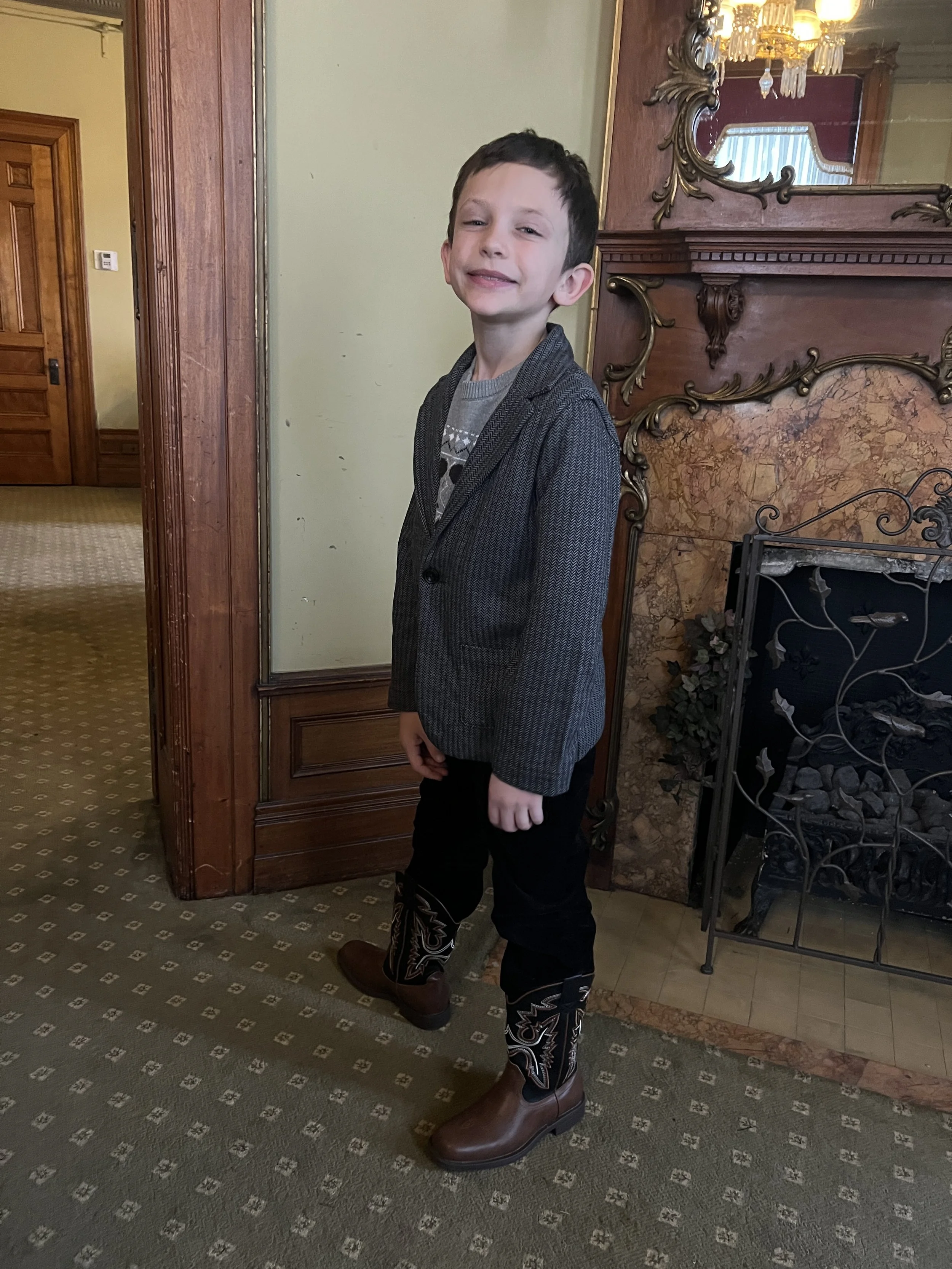 A young boy with short dark hair, wearing a gray blazer over a patterned shirt, black pants, and cowboy boots, standing in a room with vintage decor, including a wooden fireplace mantel and chandelier, smiling slightly.