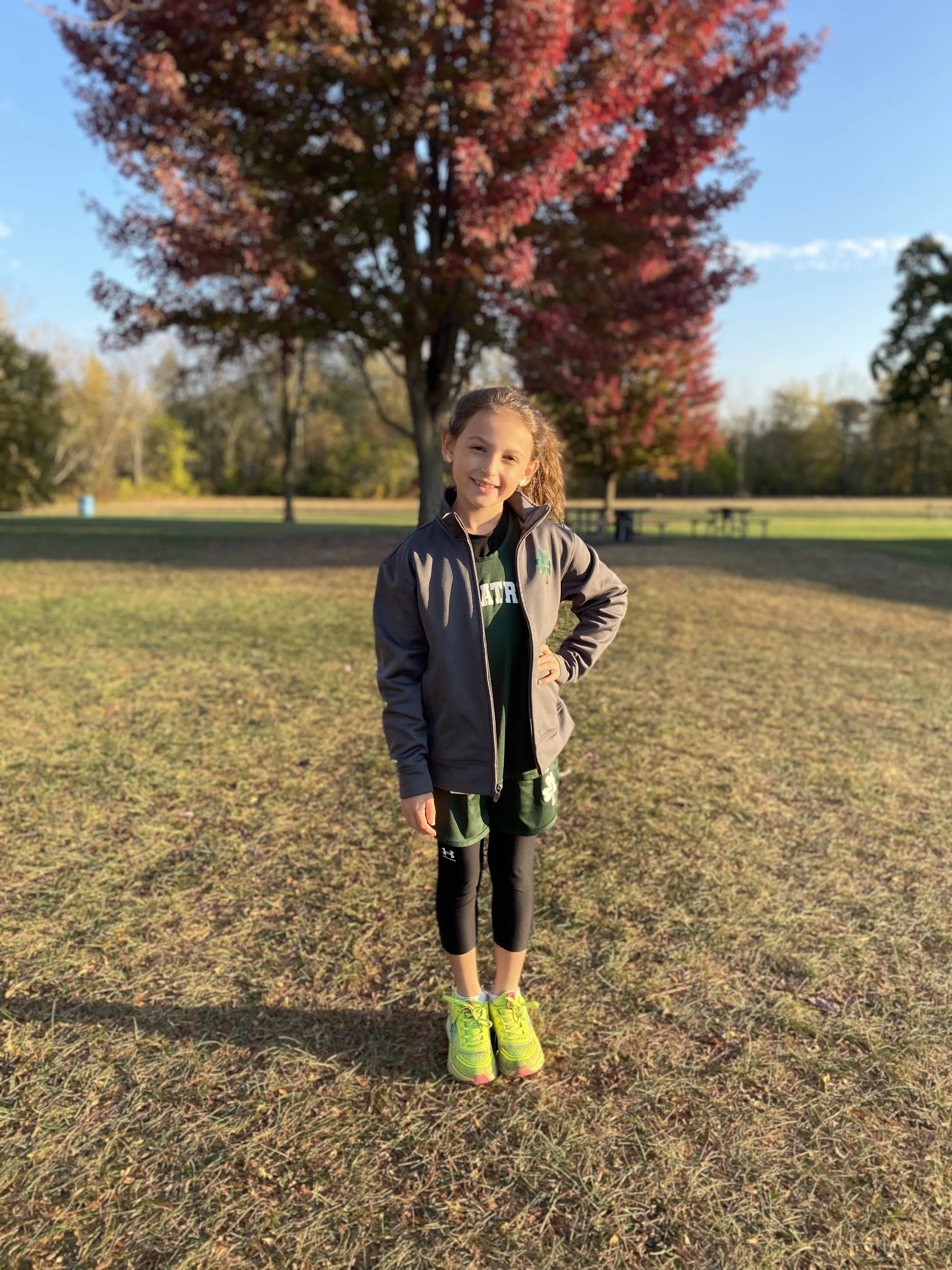 A young girl standing outdoors on a grassy field with a large red-leafed tree behind her, wearing athletic clothes and bright yellow running shoes, during a sunny autumn day.