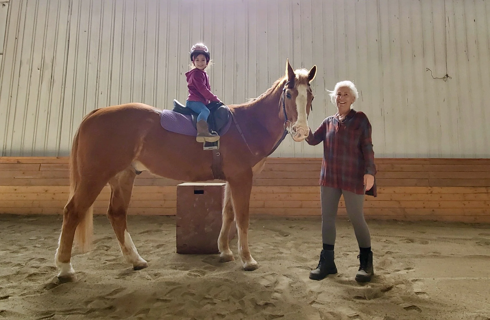 A young girl riding a horse in an indoor riding arena, with an older woman standing next to the horse holding its reins.