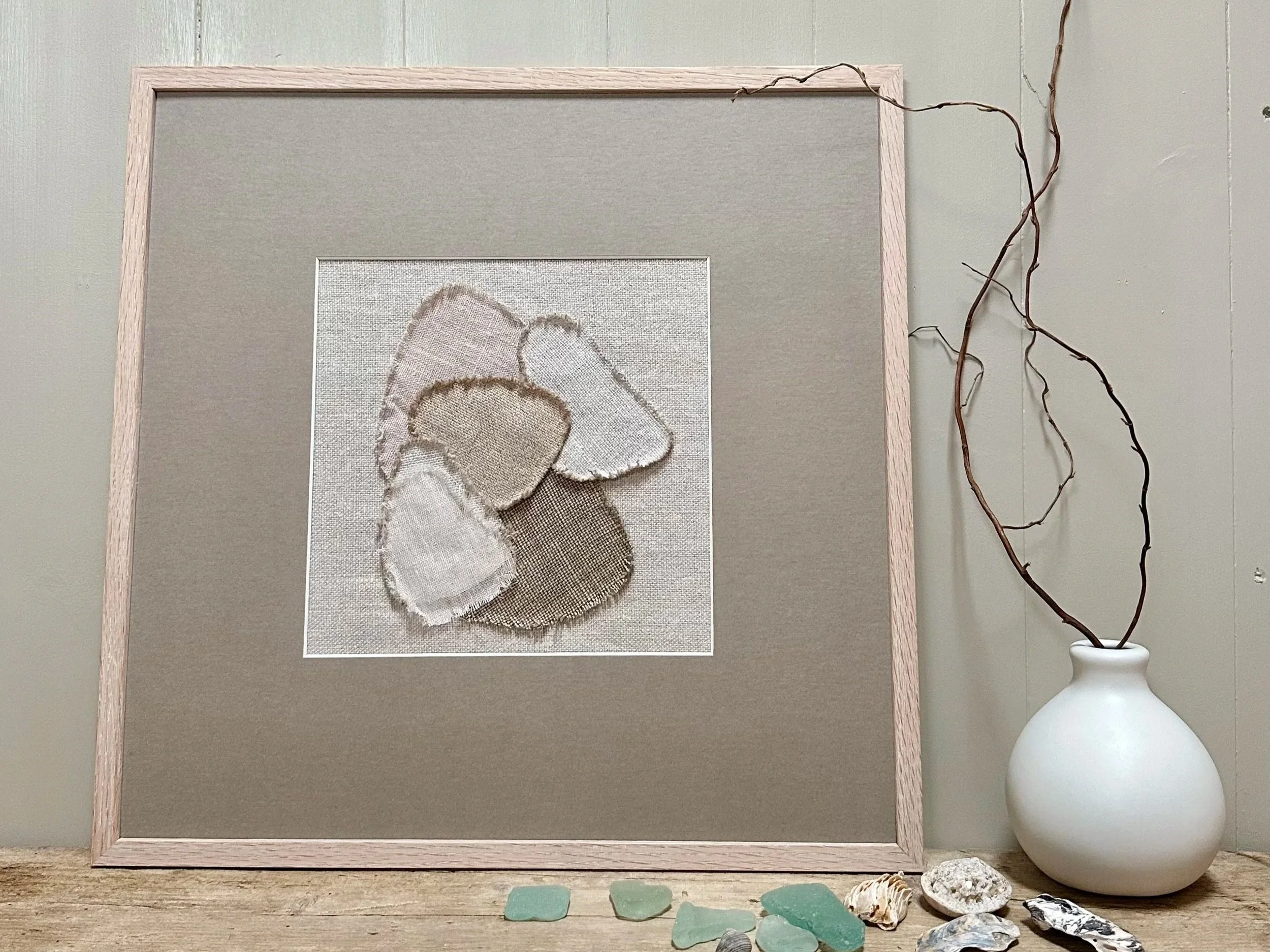 A framed embroidery of a teddy bear holding a heart sits on a wooden surface beside a white vase with twisted branches. Several small stones are scattered in front of the frame.
