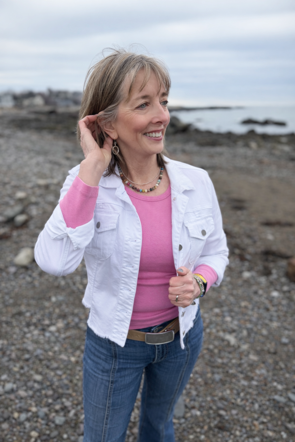 A woman with light brown hair wearing a pink shirt and white jacket stands on a rocky beach, smiling and looking to the side. The background shows cloudy skies and a shoreline with rocks and water.